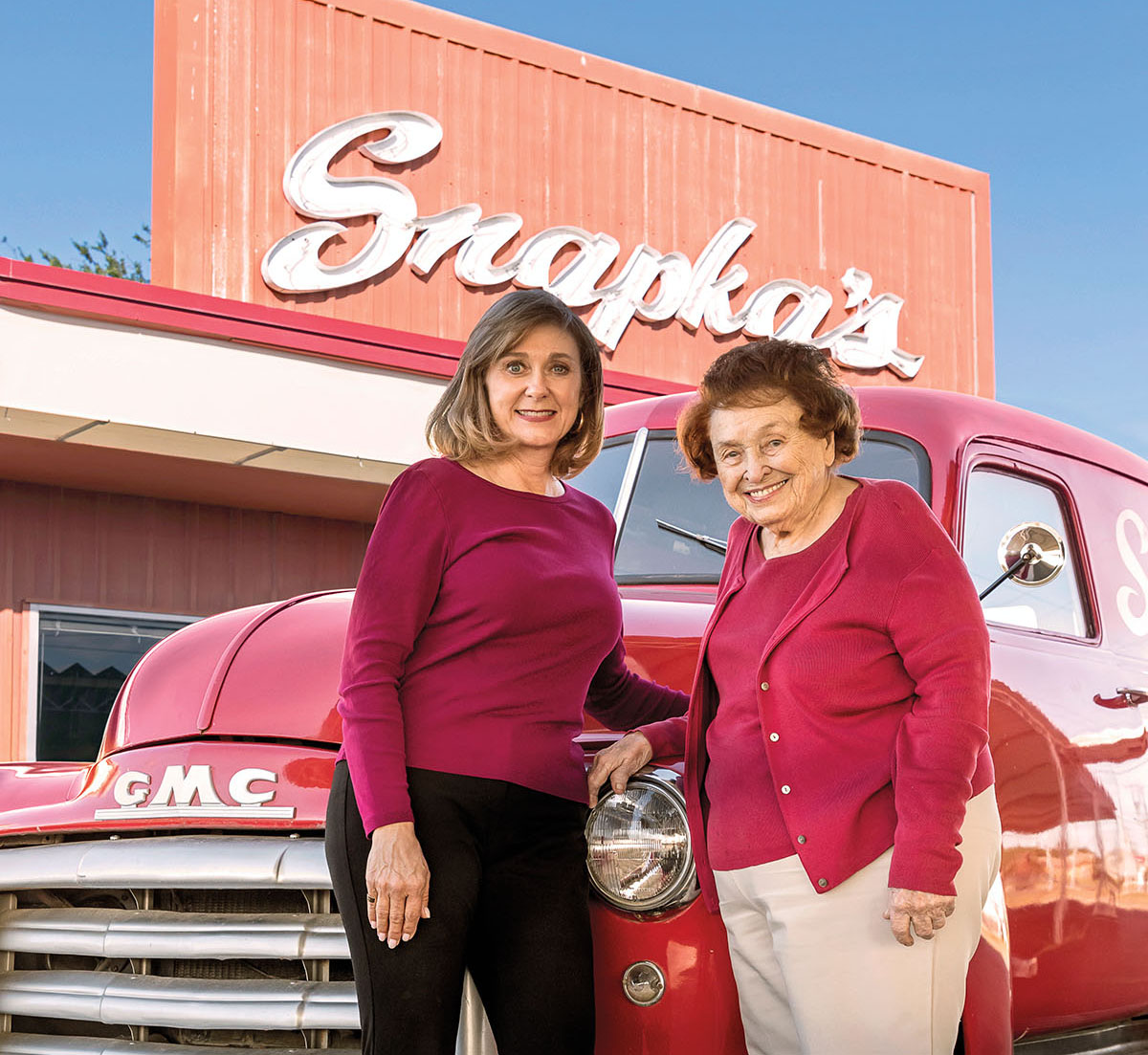 Two women in red shirts stand in front of a bright red GMC truck and a sign reading "Snapka's"