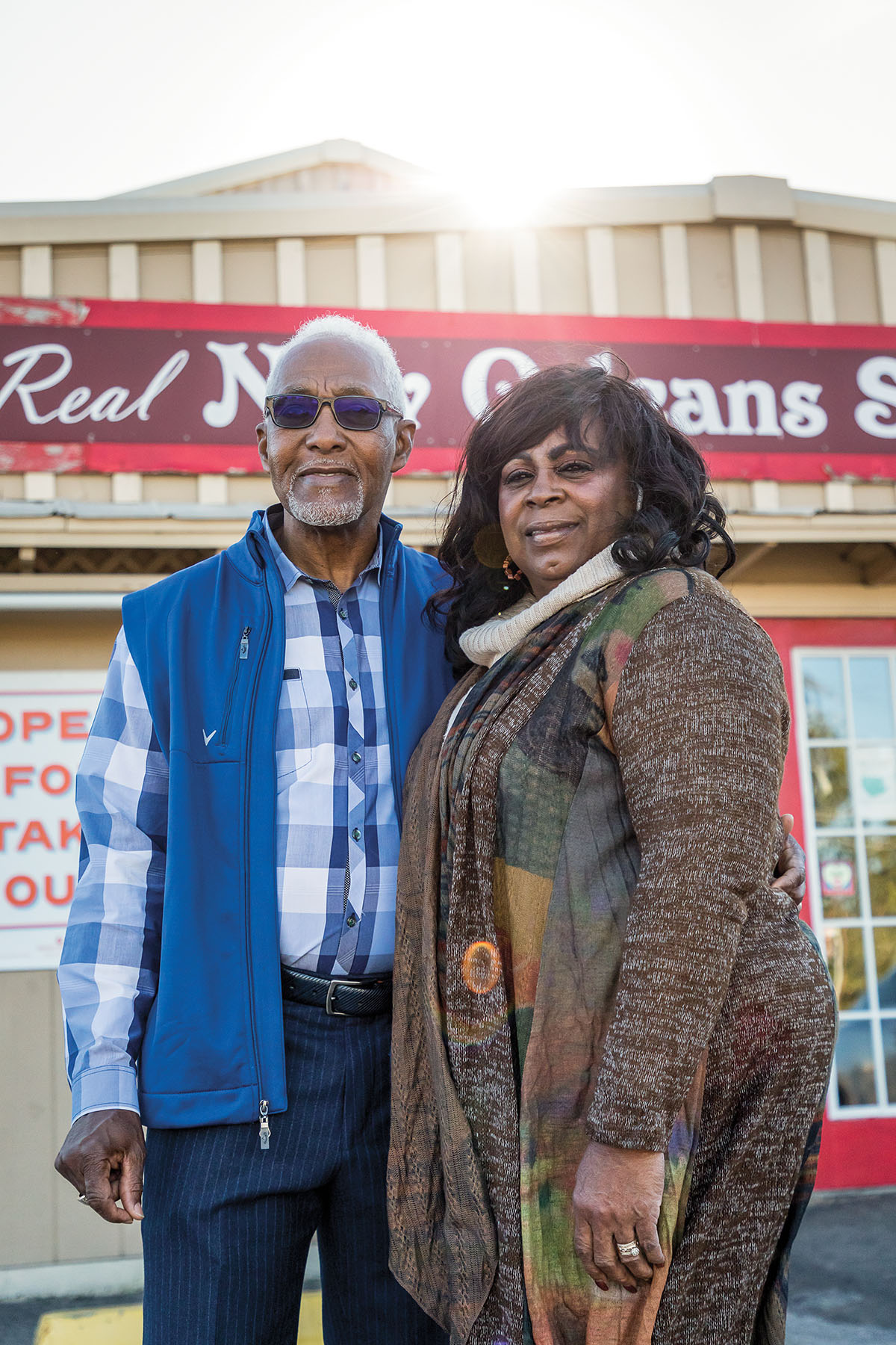 A man in a blue shirt and vest and a woman in a heathered brown shirt stand outside of the restaurant