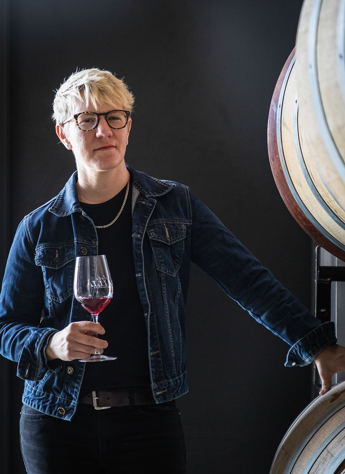 A person in a denim jacket and black shirt stands with a glass of wine in hand next to a wine barrel