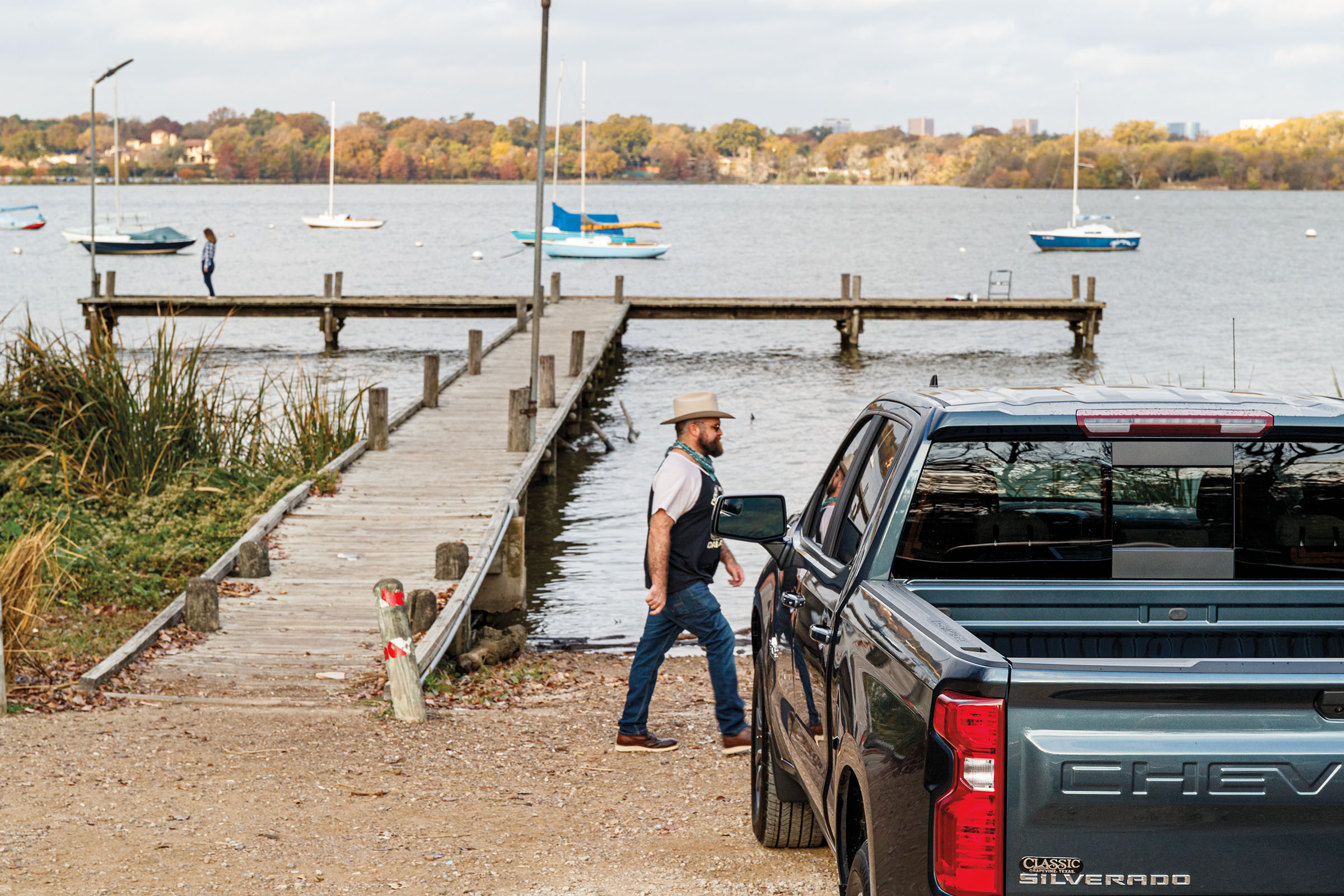 A dark teal pickup truck in the foreground of a photo with a long dock, boats, and a man in overalls walking by