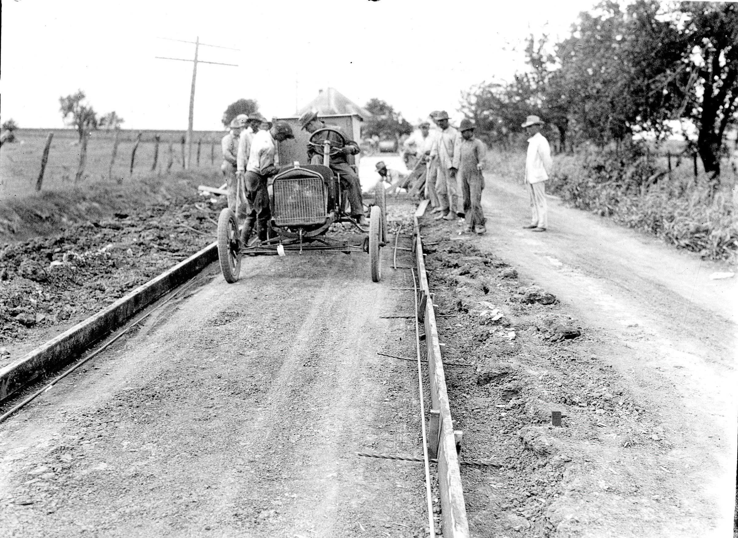 A group of men, some in an old car and some standing, lay dirt and boards