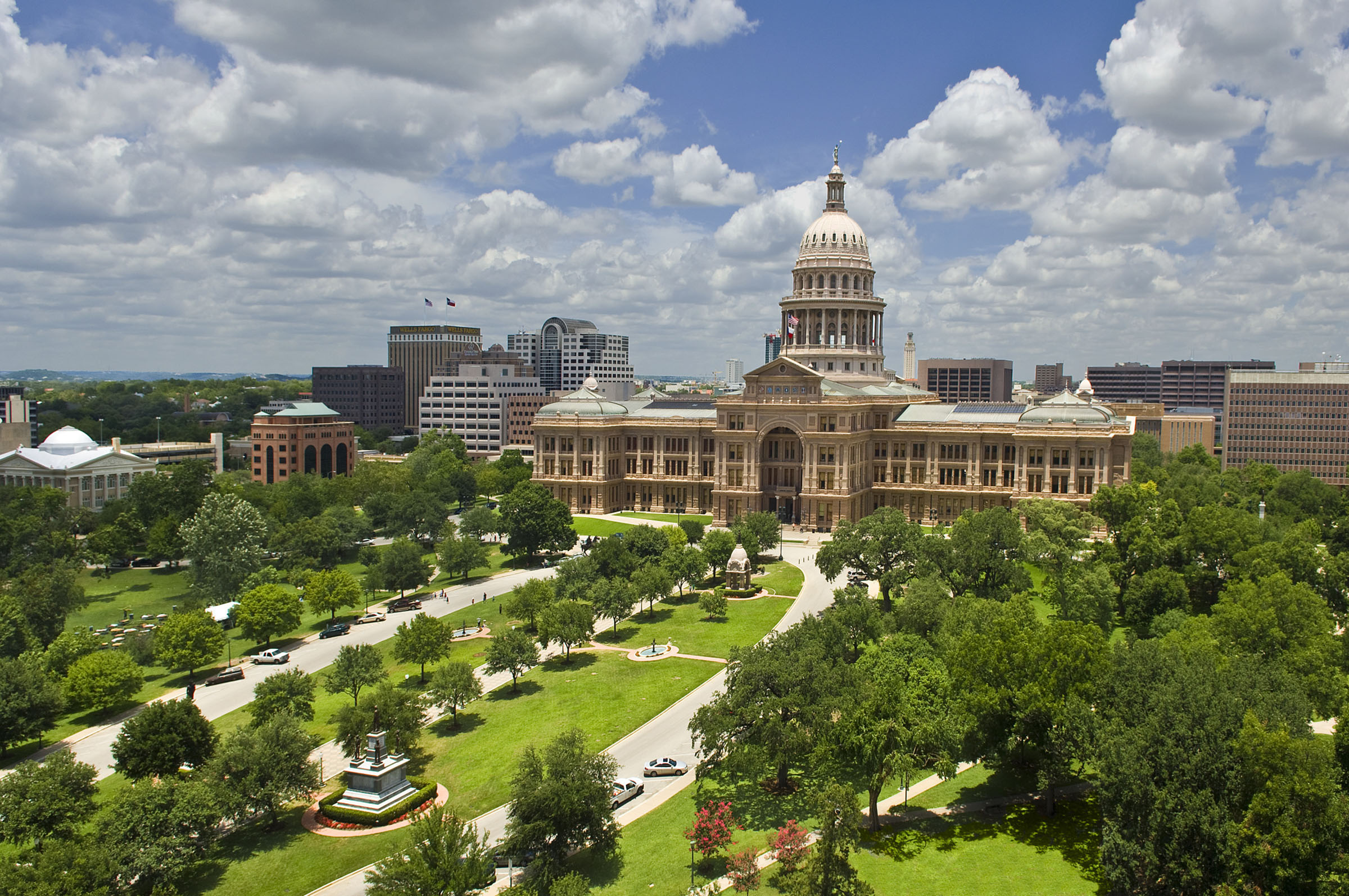 Lush green trees and grass line the driveways and sidewalks leading toward the capitol building in downtown Austin, Texas. The UT Tower is also visible in the background