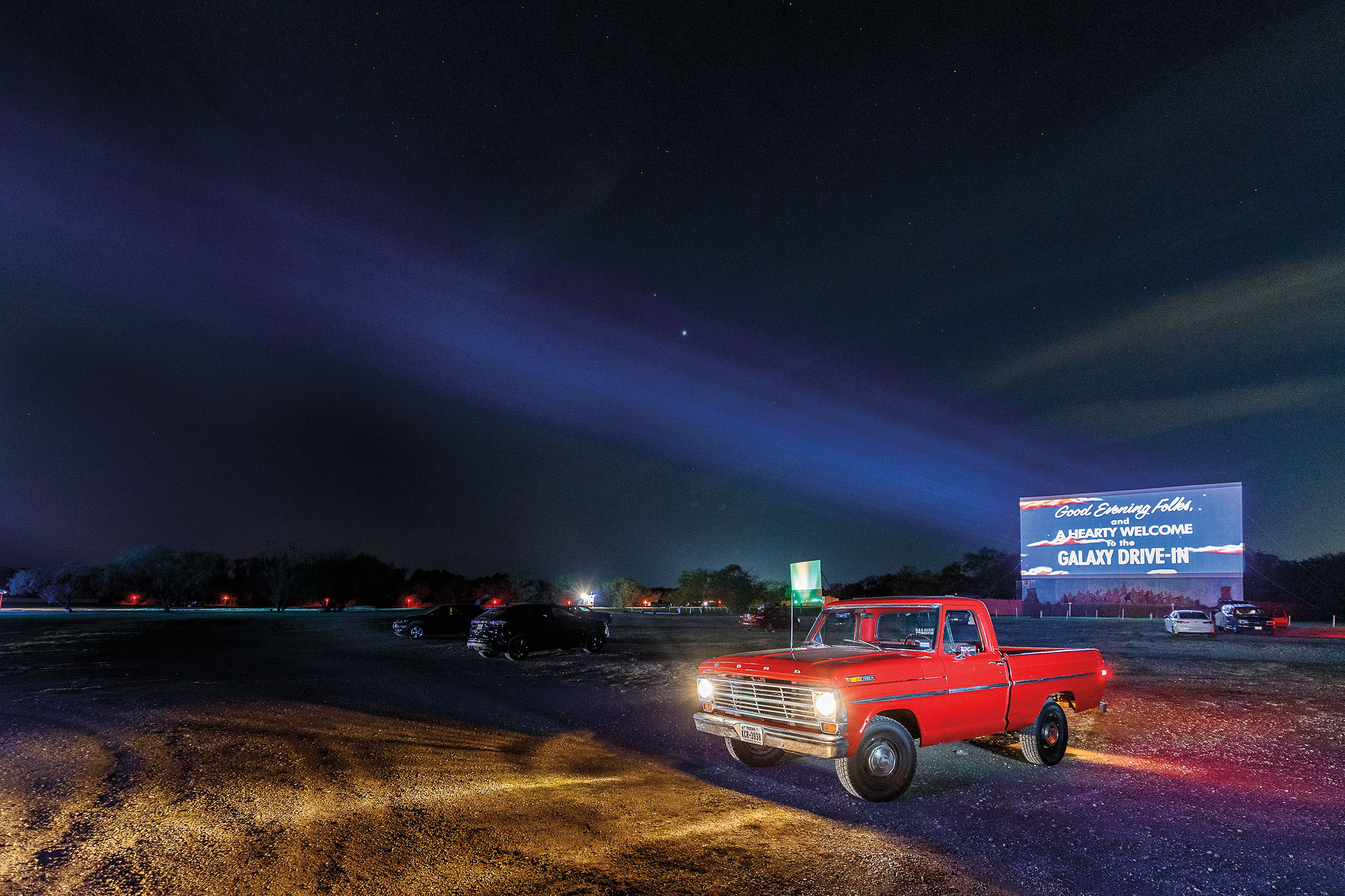 A red ford truck with the headlights on parked at the Galaxy Drive-In, with the large blue movie screen visible in the background