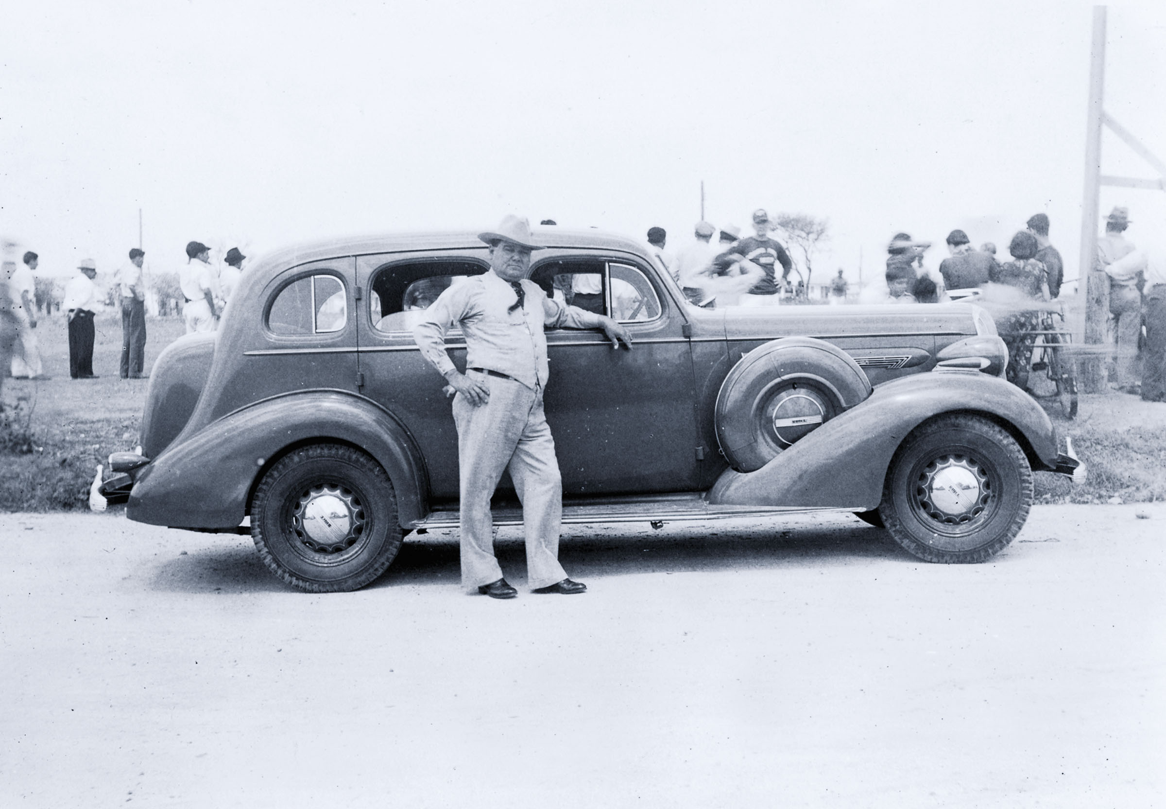 A man in a short cowboy hat, shirt and tie leans his arm against the passenger door of a Buick Roadster