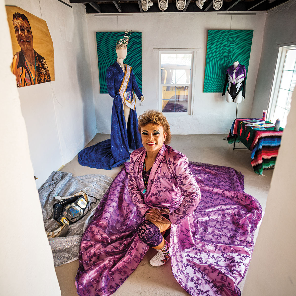 A person in a large, flowing purple gown sits on the floor in a white room with large paintings on the wall
