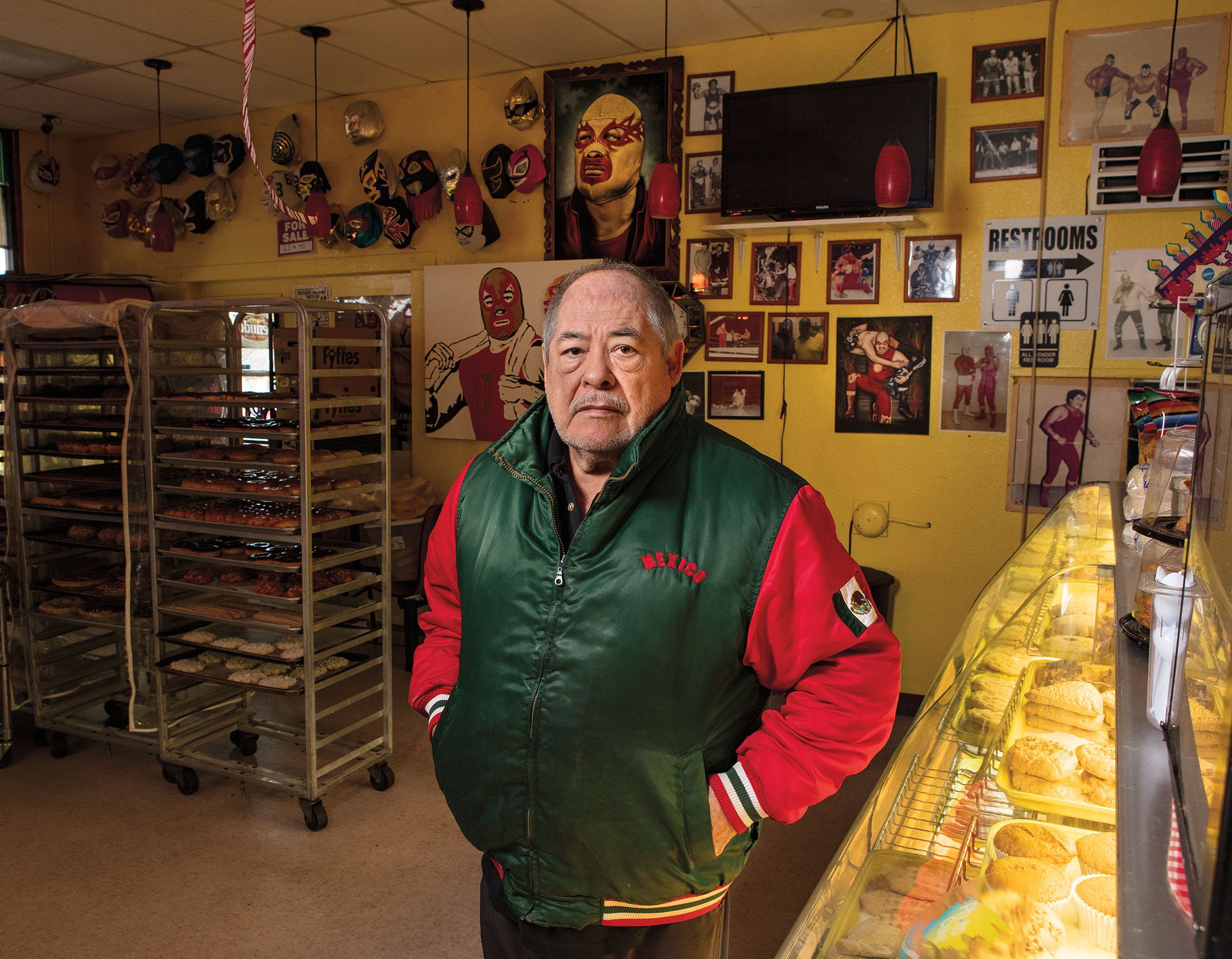 A man in a green and red jacket stands in a bakery, with walls filled with photos and memorabilia of lucha libre
