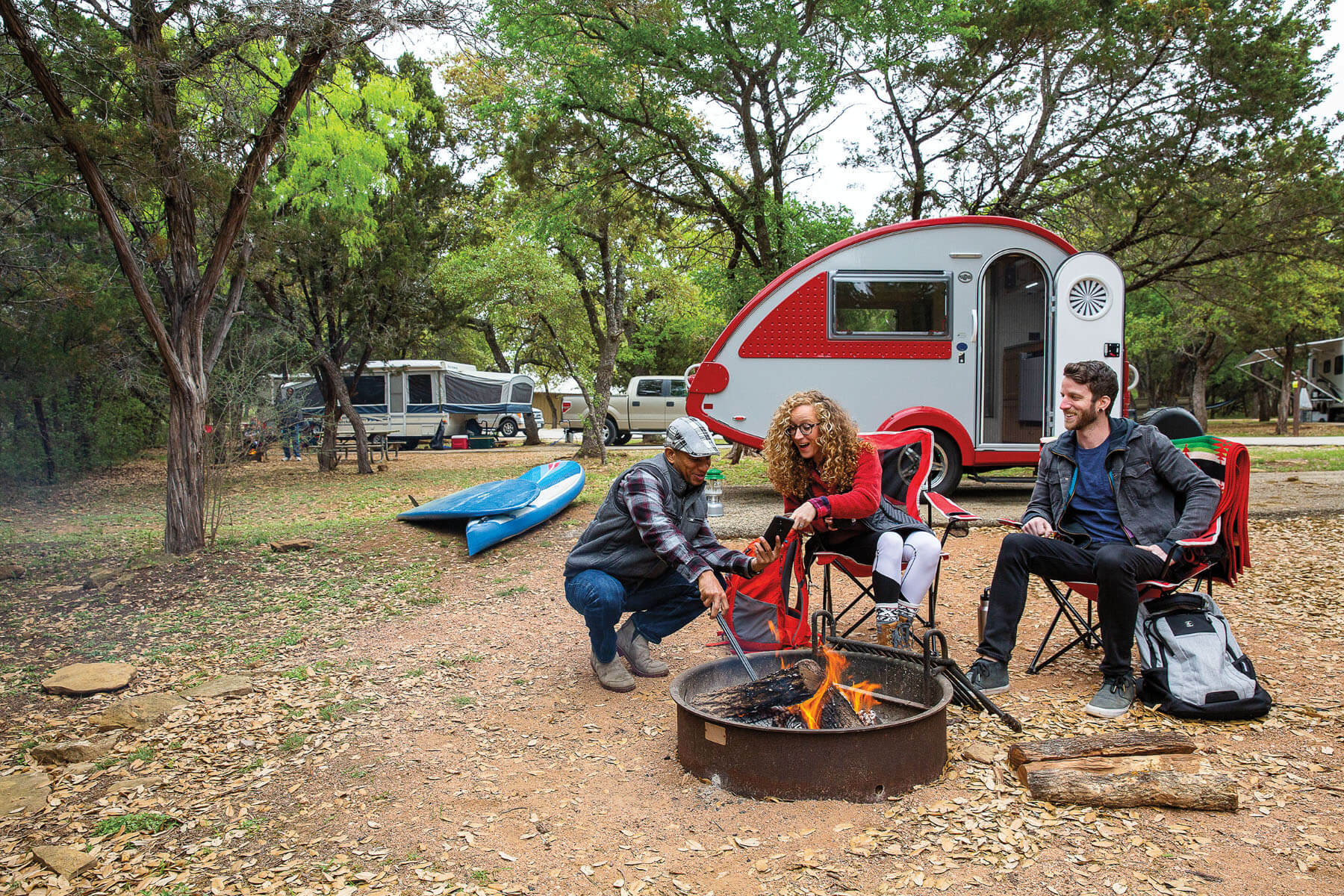 A group of people sit in front of a red, teardrop-shaped camper around a campfire in a wooded setting