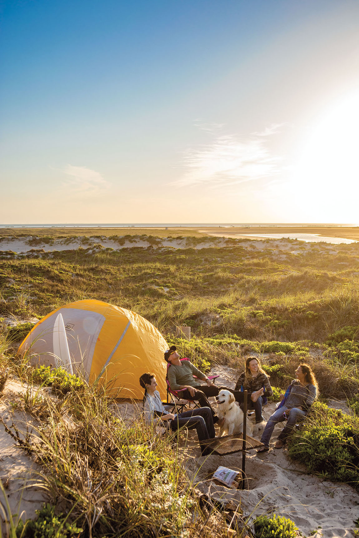 A group of people sit in the sand in camp chairs next to an orange tent on some sand dunes