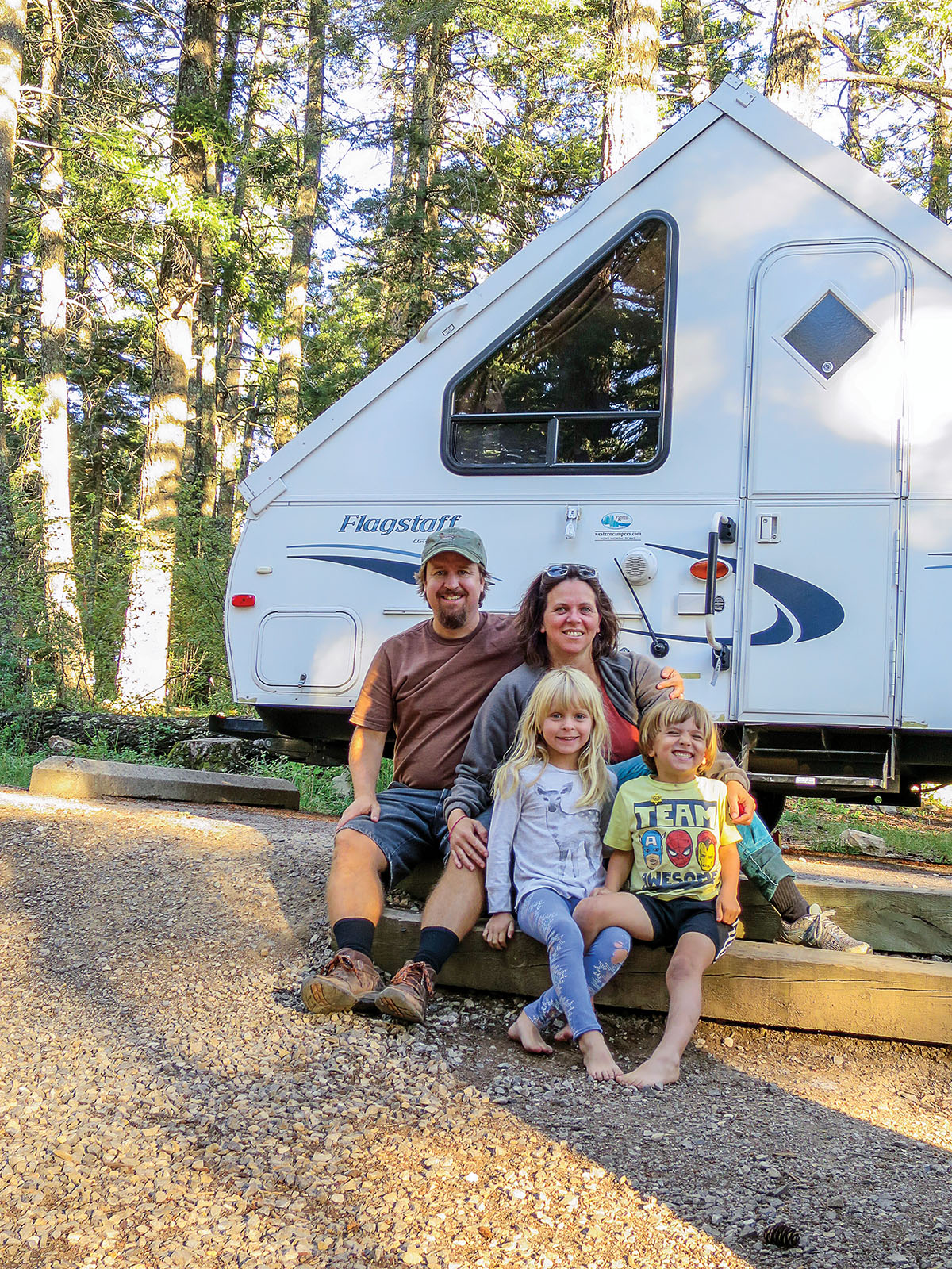 A family sits in front of a small, white, A-frame camper in a forest with tall trees