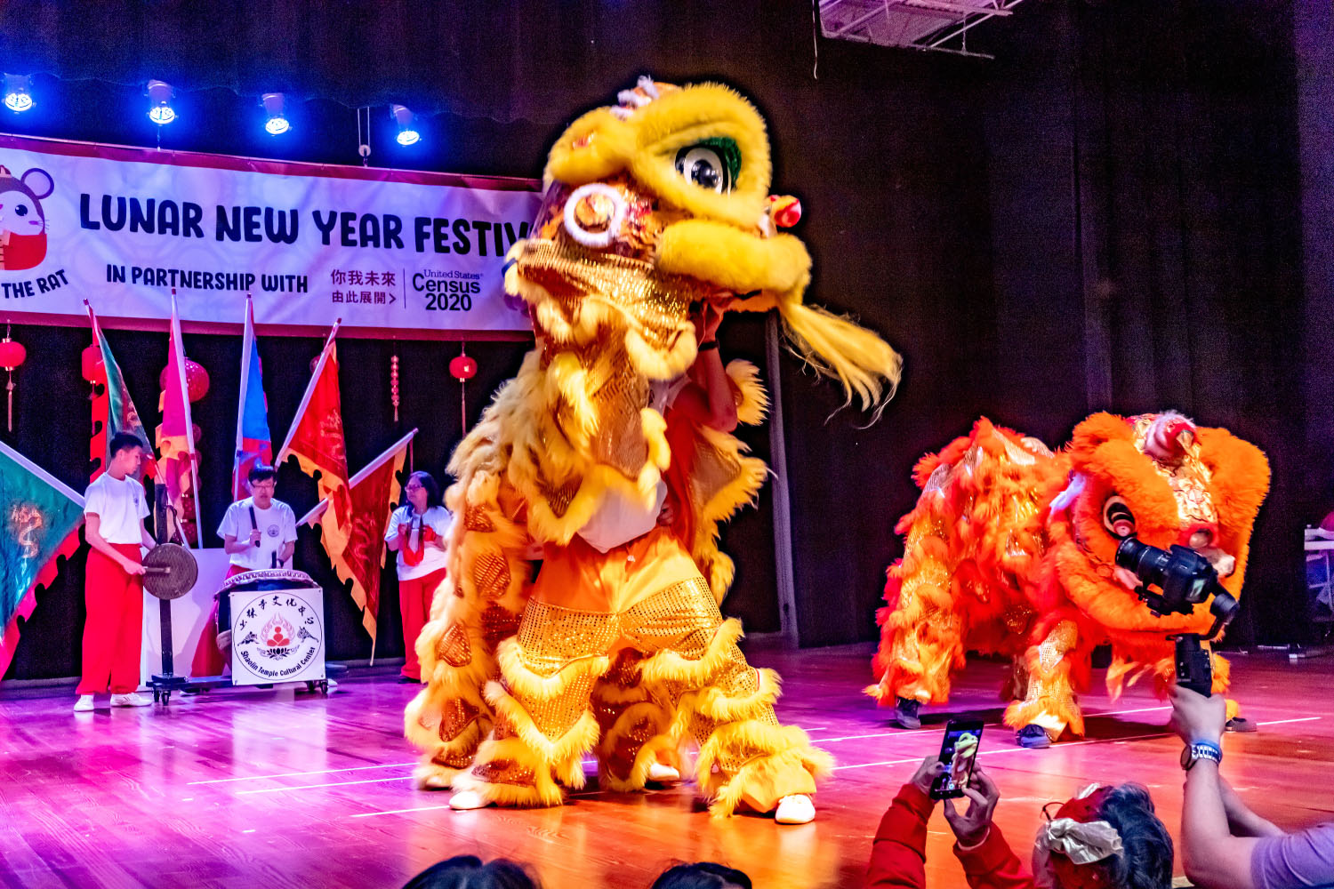 Brightly-colored costumed dragons perform on a stage in front of drummers and flags