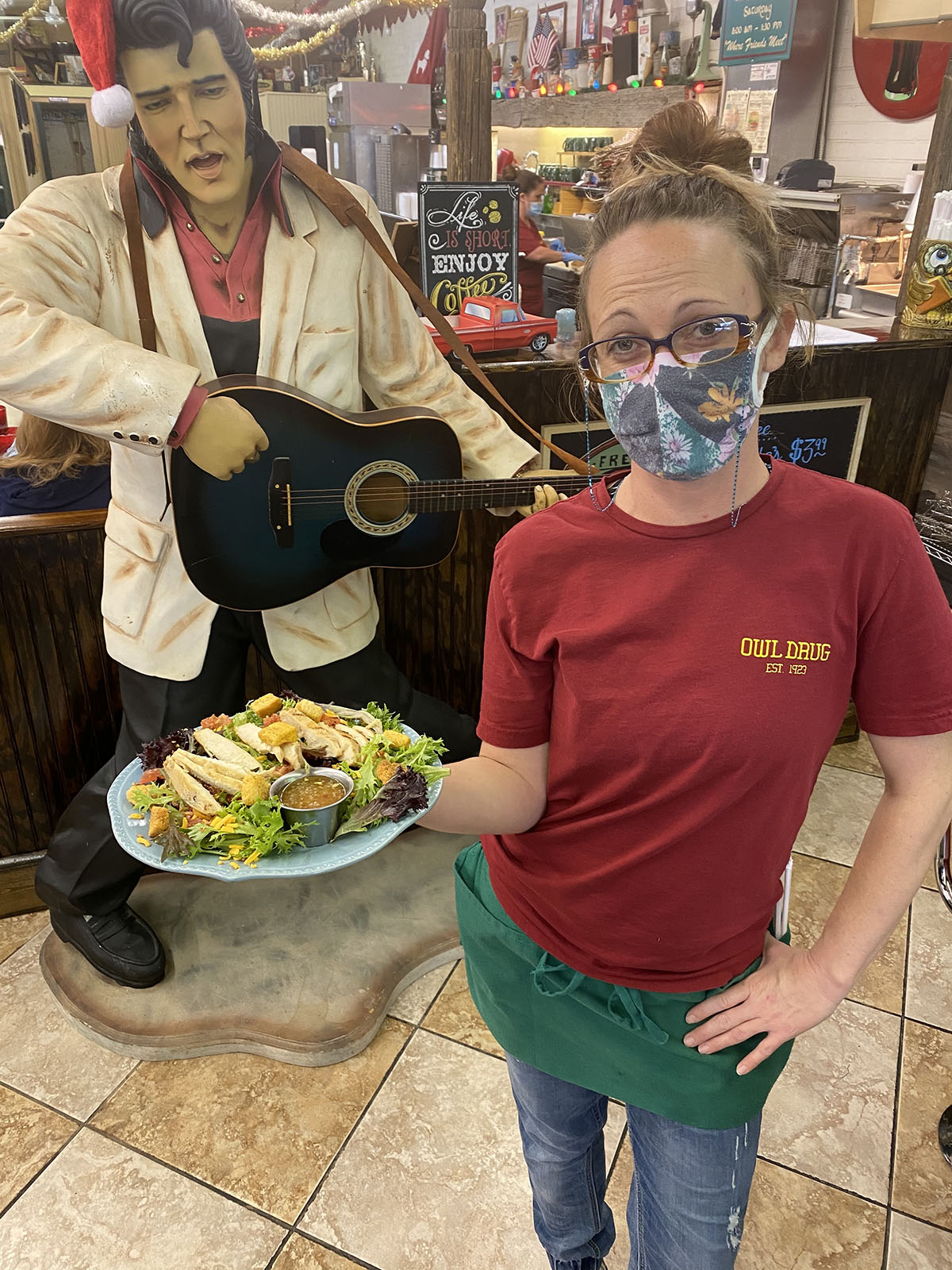 A waitress hold a plate with a large salad and stands in front of the life-size Elvis statue at Owl Drug.