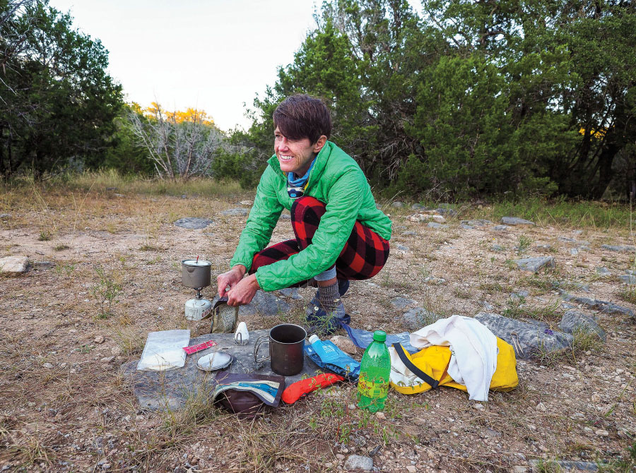 A woman in a green long-sleeved shirt stands among backpacking equipment
