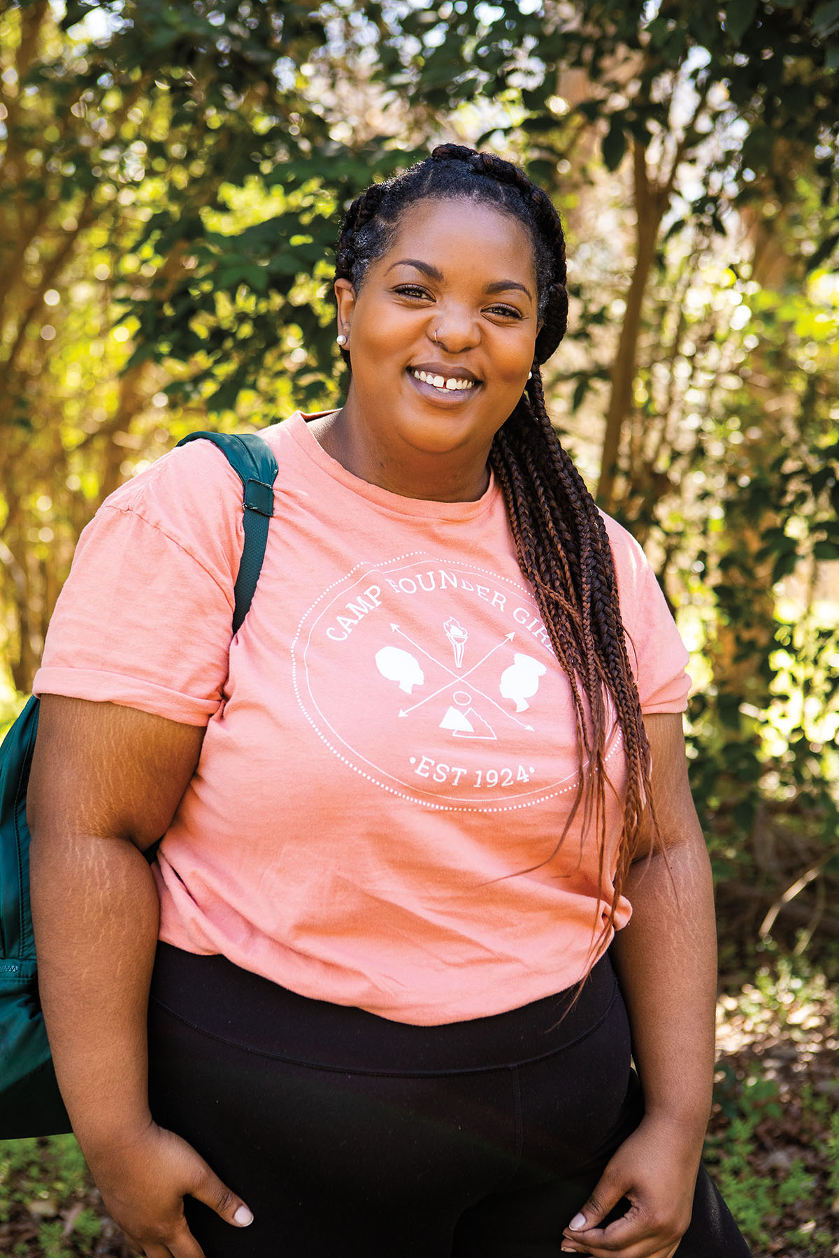 A woman in a pink t-shirt wearing a backpack stands in a tree-filled setting