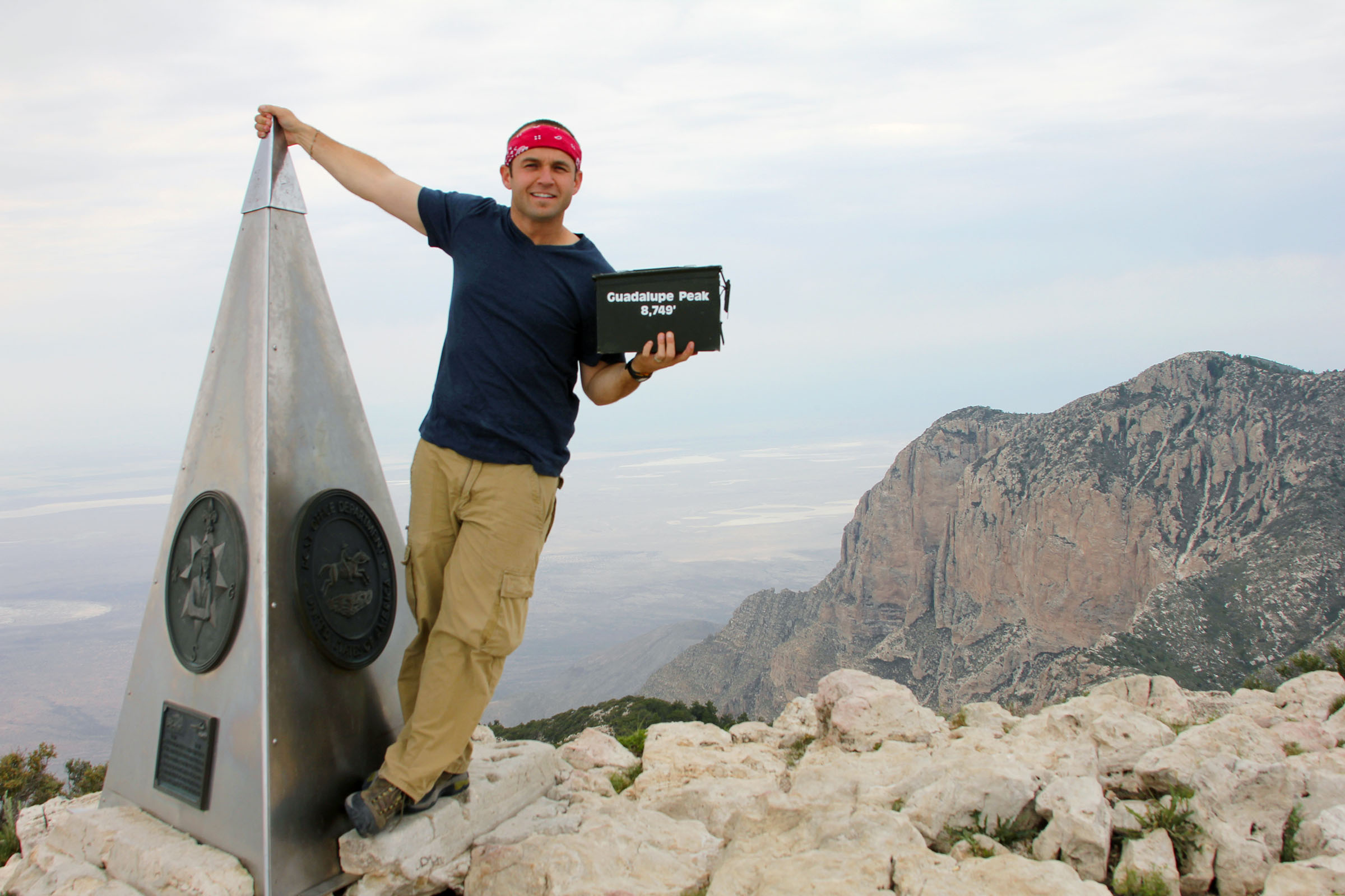 A man in a black t-shirt and red bandana leans on the "top of Texas" marker on Guadalupe Peak