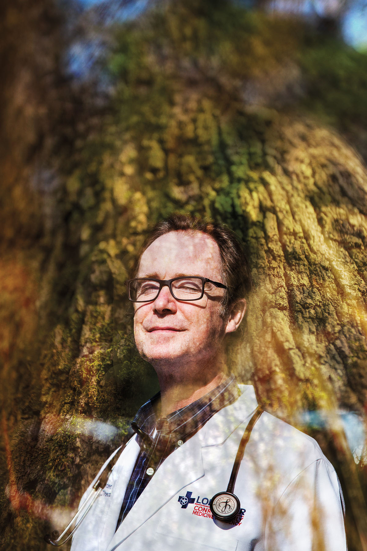 A man in a doctor's overcoat and stethascope stands double-exposed with a large tree