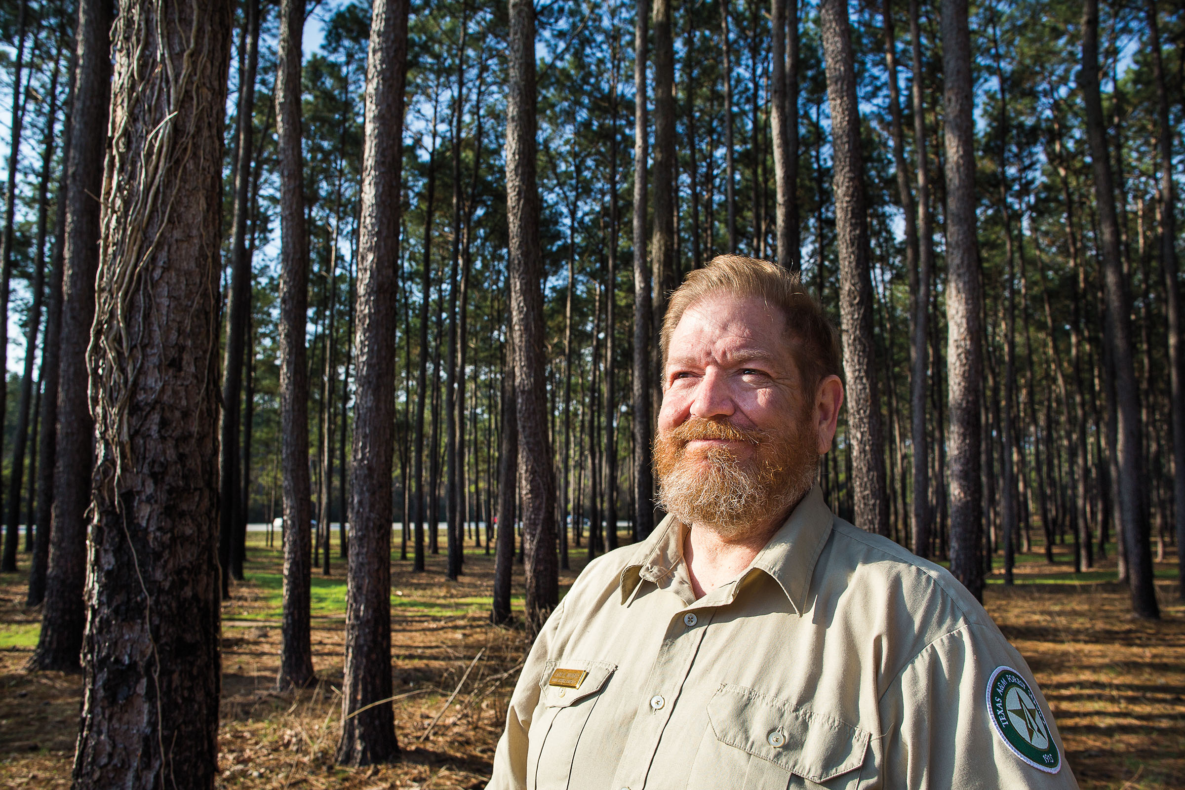 A man with a beard in a Forest Service shirt stands in a grove of trees