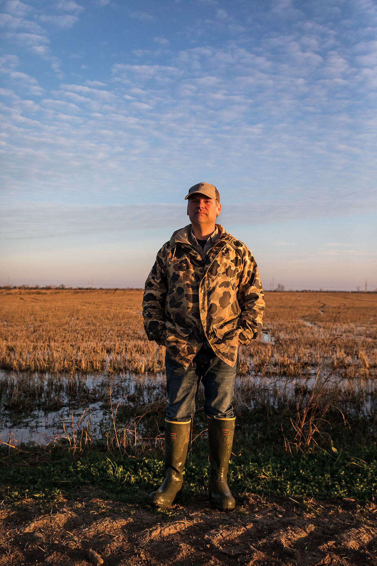 A man in a camoflague jacket and boots stands with his hands in his pockets in a bayou