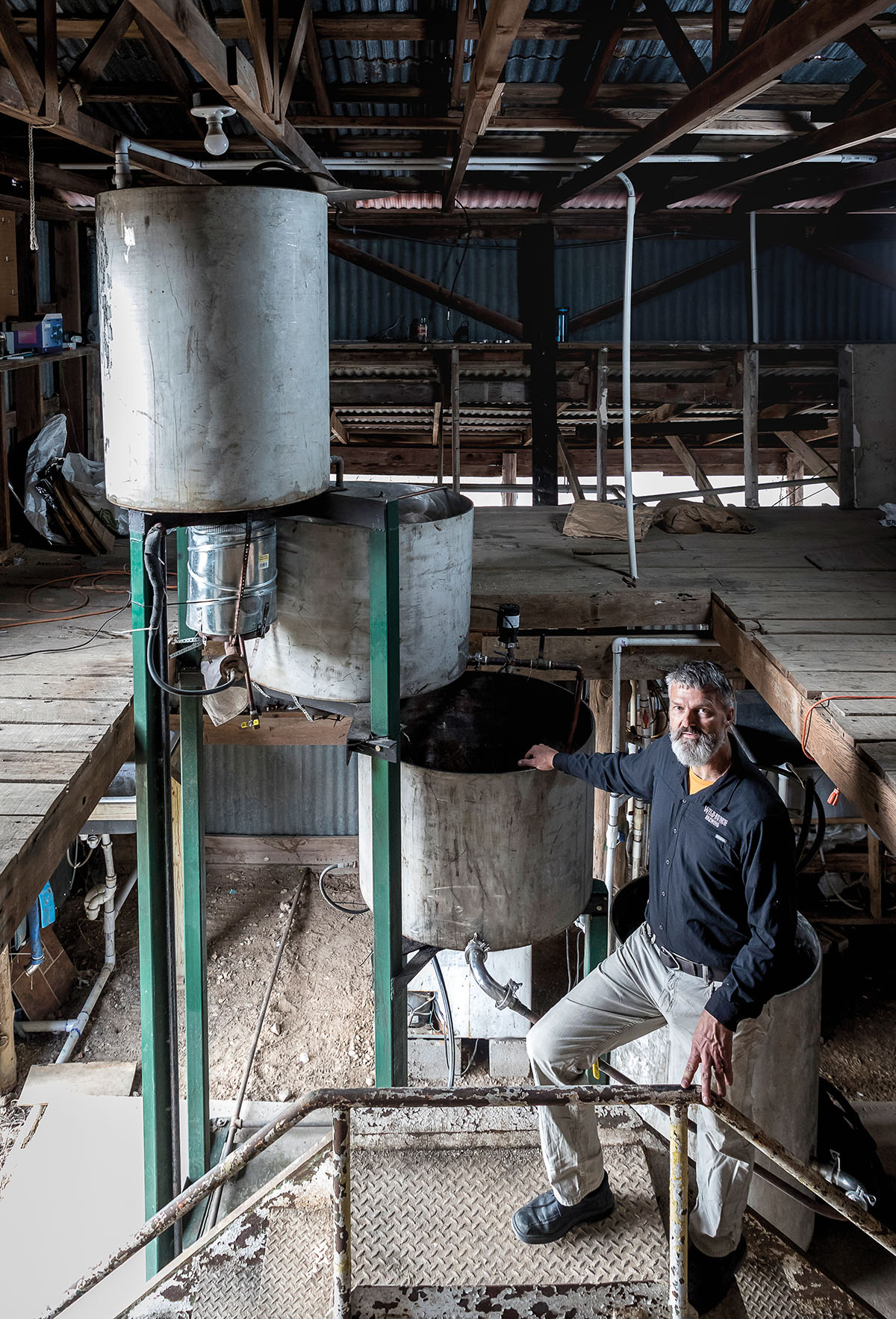 A man in a blue shirts stands next to several large metal pots in a wooden building