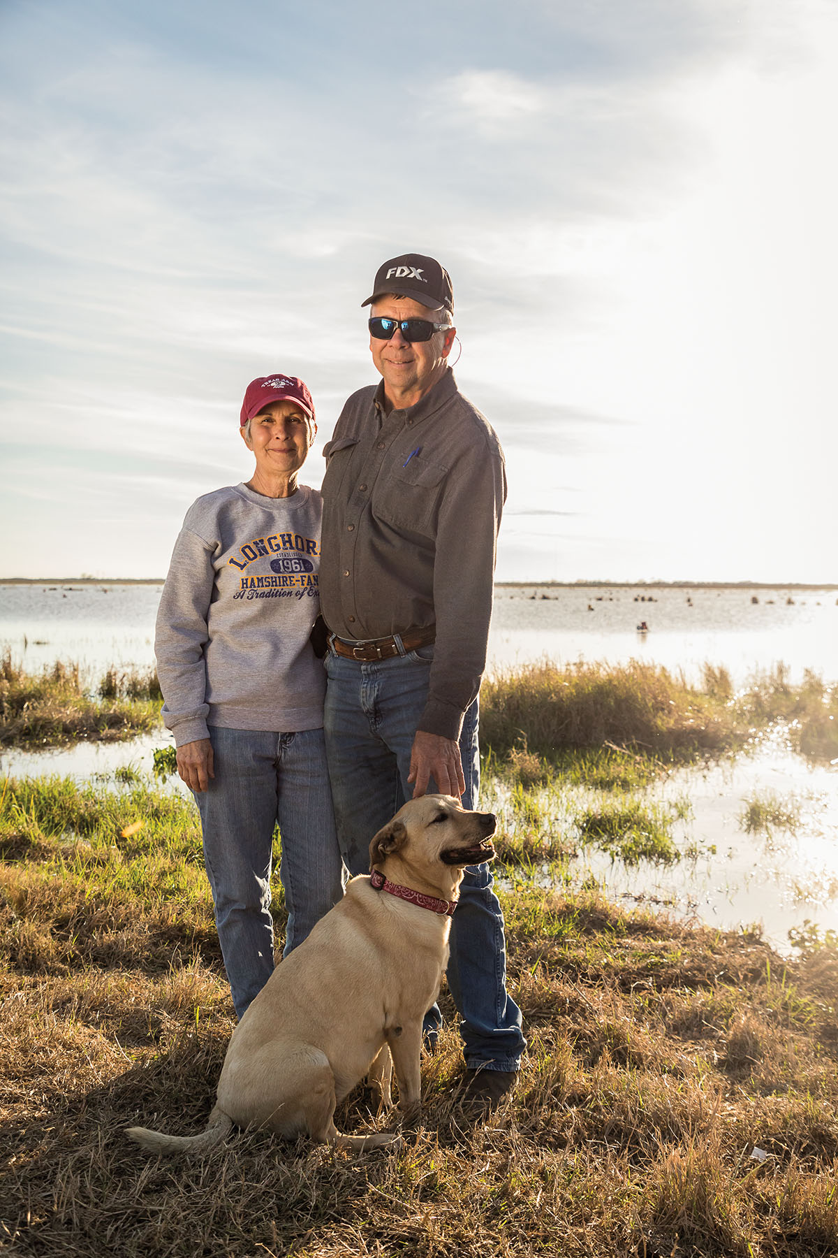 A man and woman stand with a dog on the edge of a shoreline