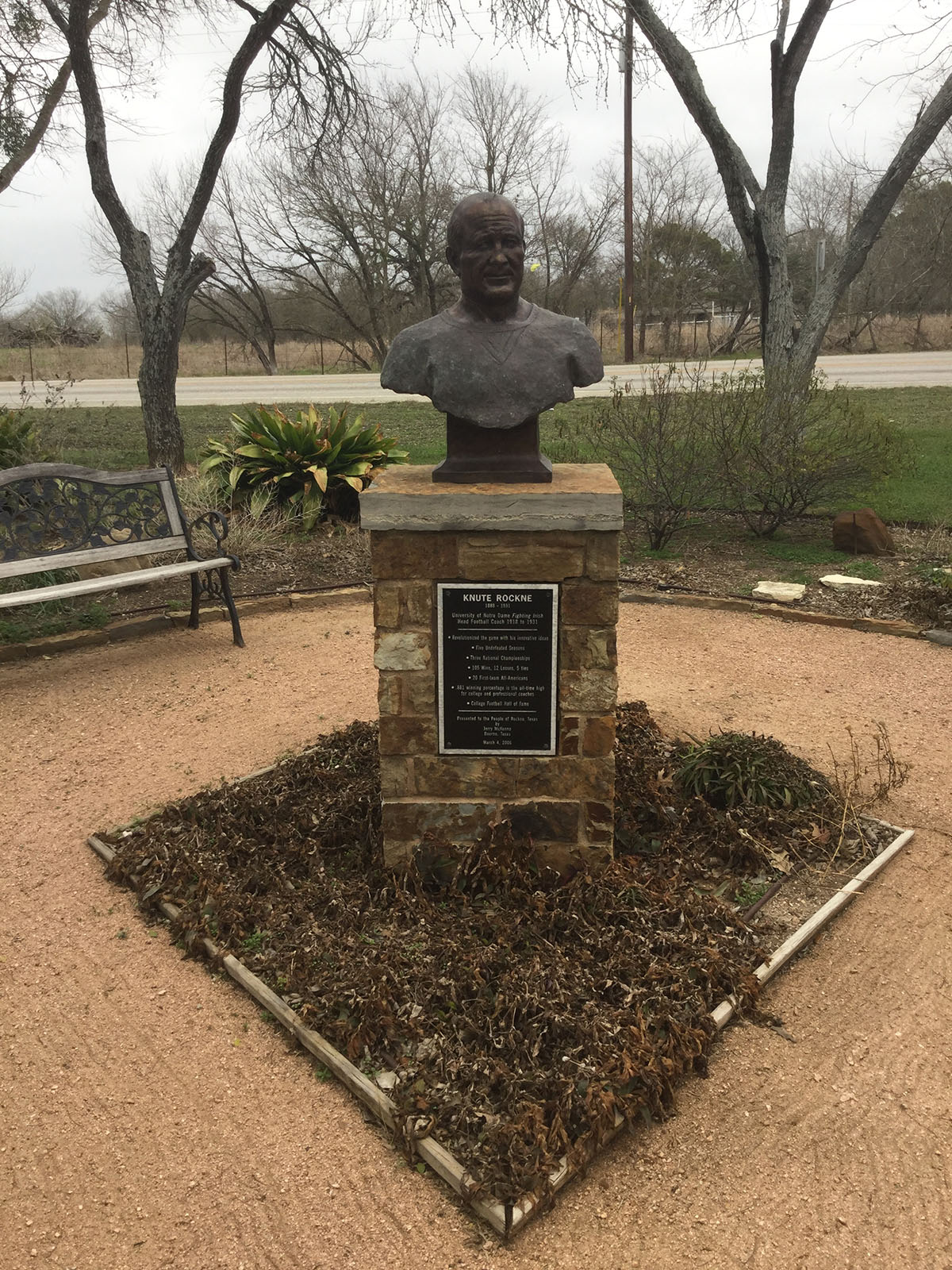 A bronze bust of Notre Dame football coach Knute Rockne sits upon a stone pedestal situated in a garden. Photo by Michael Corcoran. 