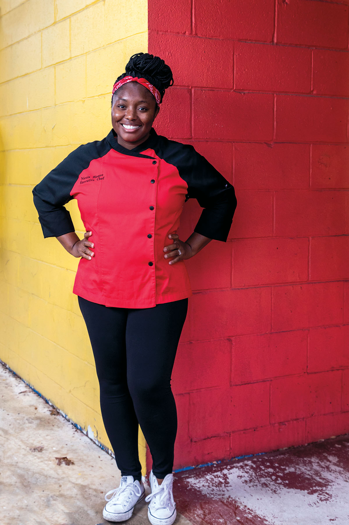 A woman in a red and black chef's jacket stands in front of a red and yellow brick wall