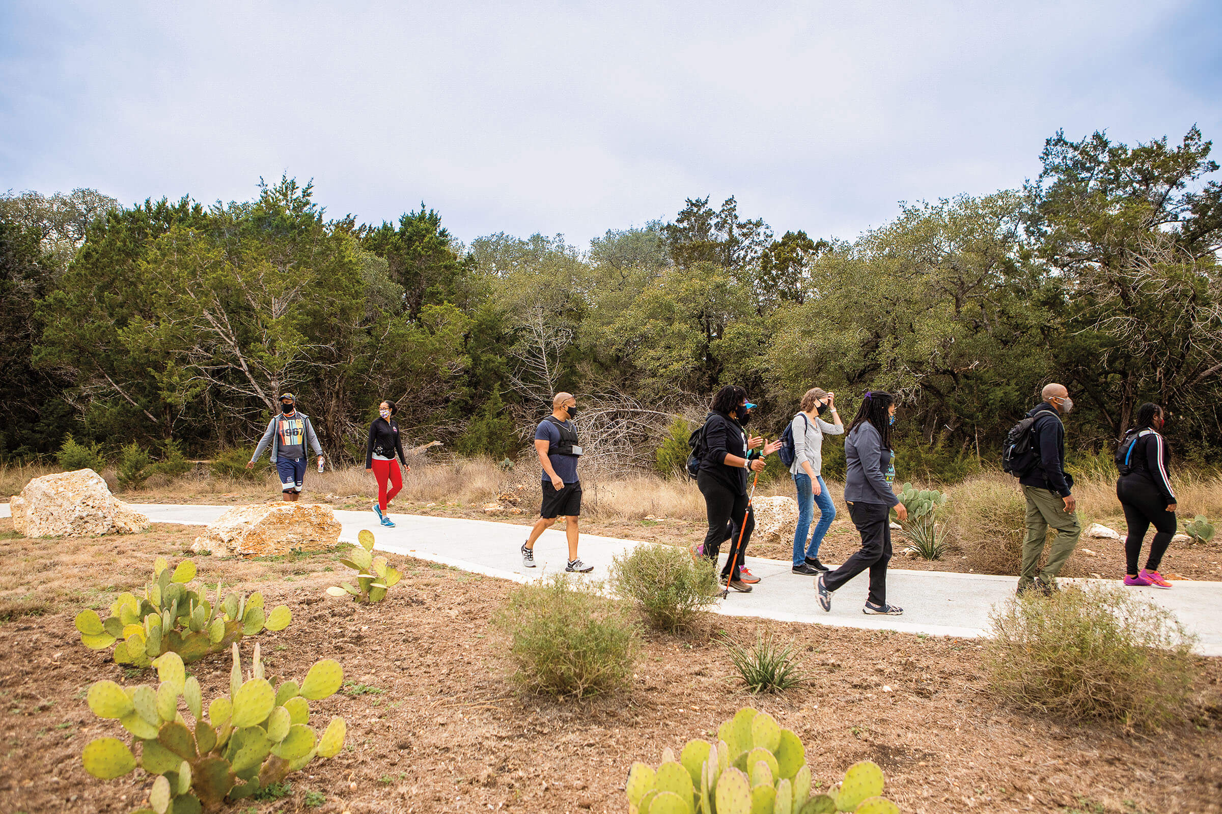 A group of people hike on a concrete trail among short brown brush and taller green trees