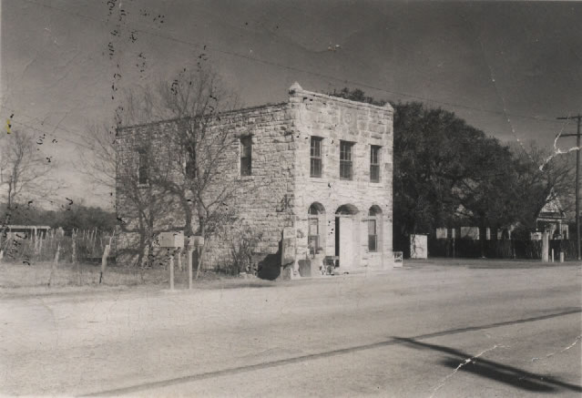 A black and white photo shows an old stone building, known as the "Old Rock Store" building, sits along a highway. 