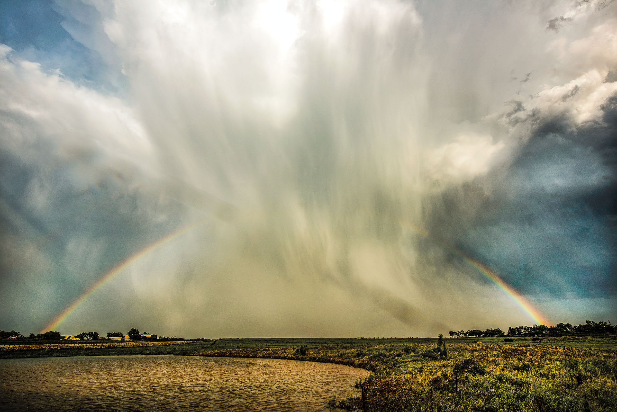 Bright white and gray clouds dump rain onto a desert landscape. A rainbow forms in the background.