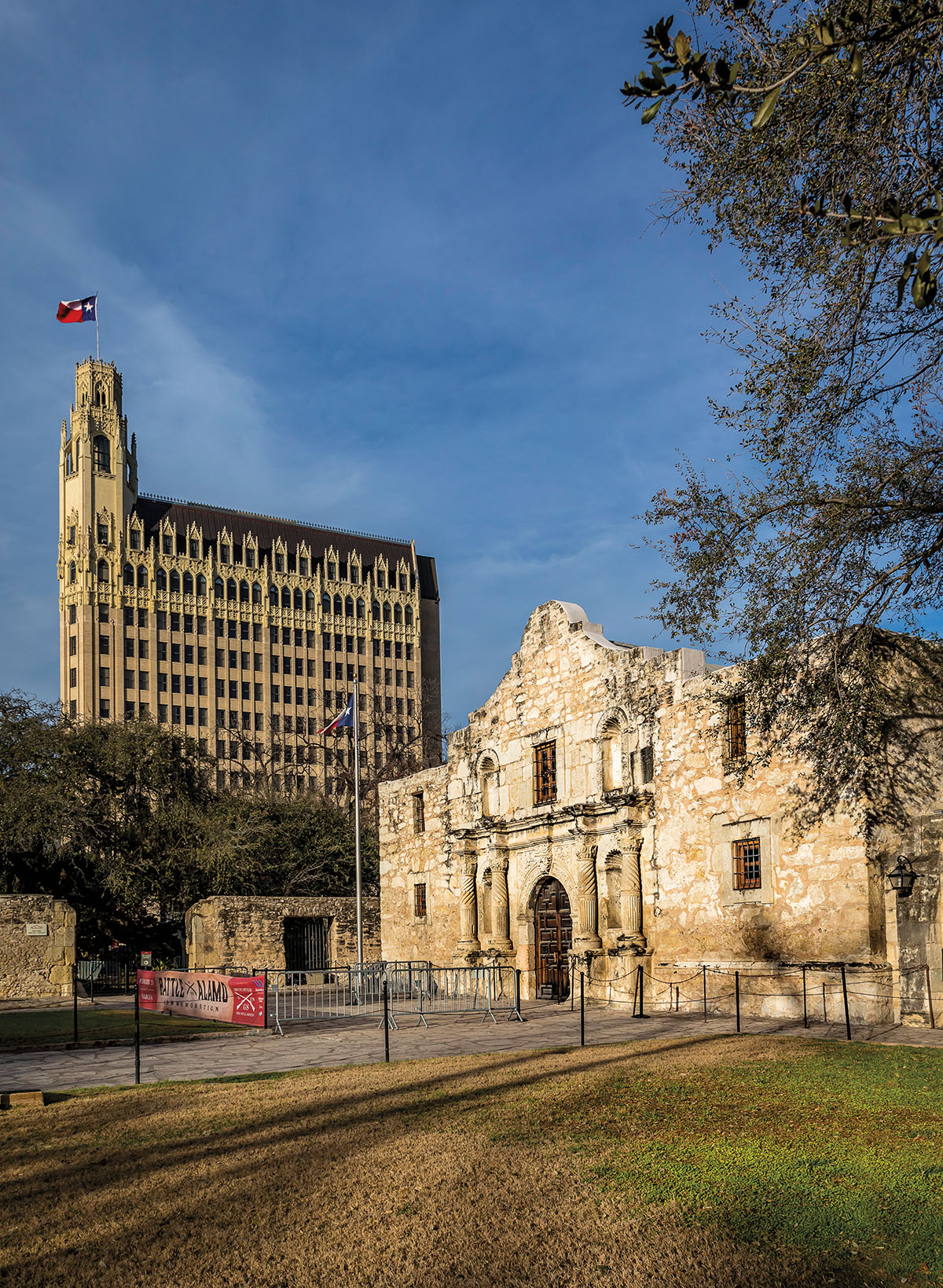 The stone facade of the Alamo in front of a taller building in the background