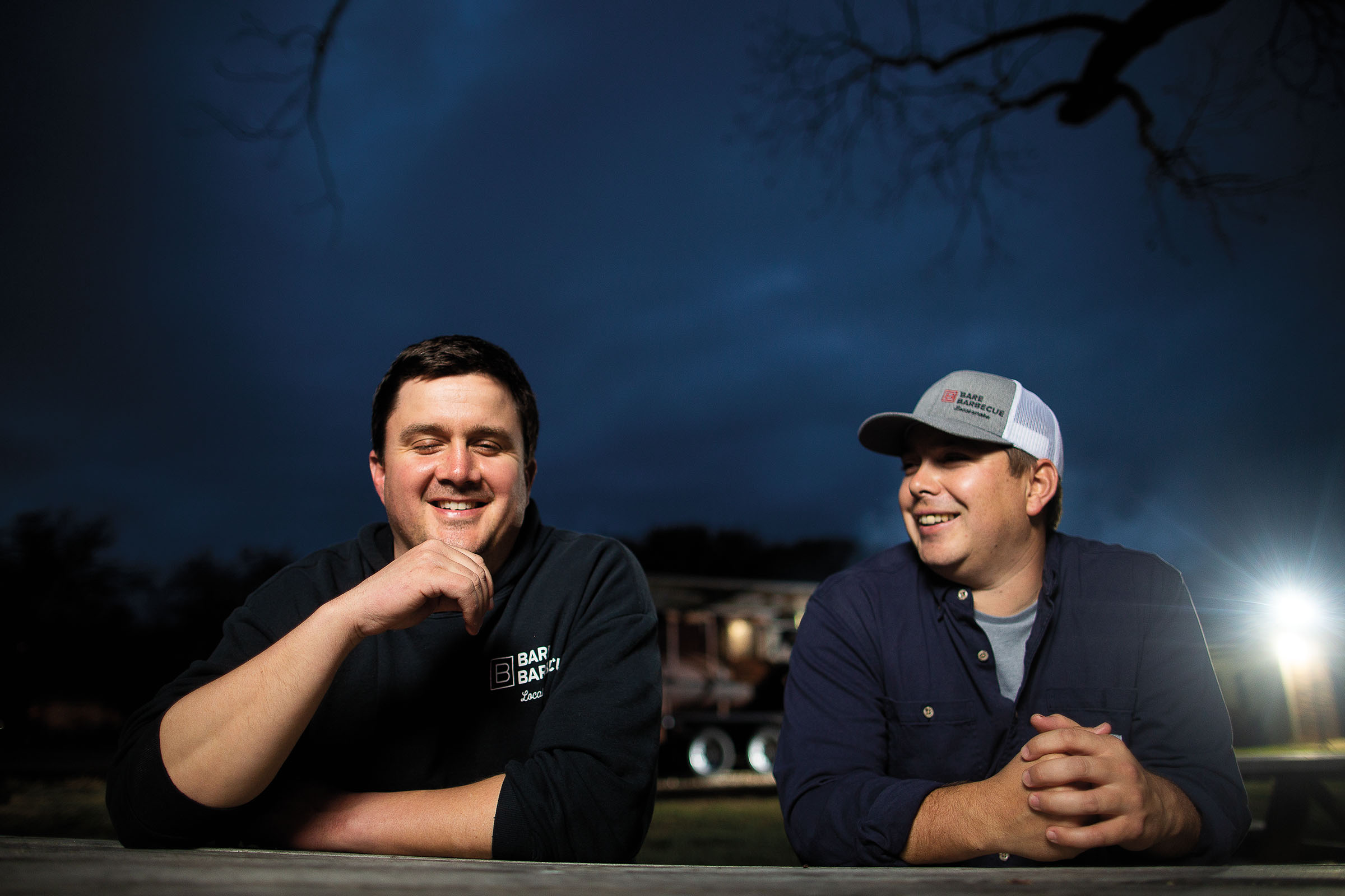 Two men laugh at a picnic table underneath a dark blue night sky