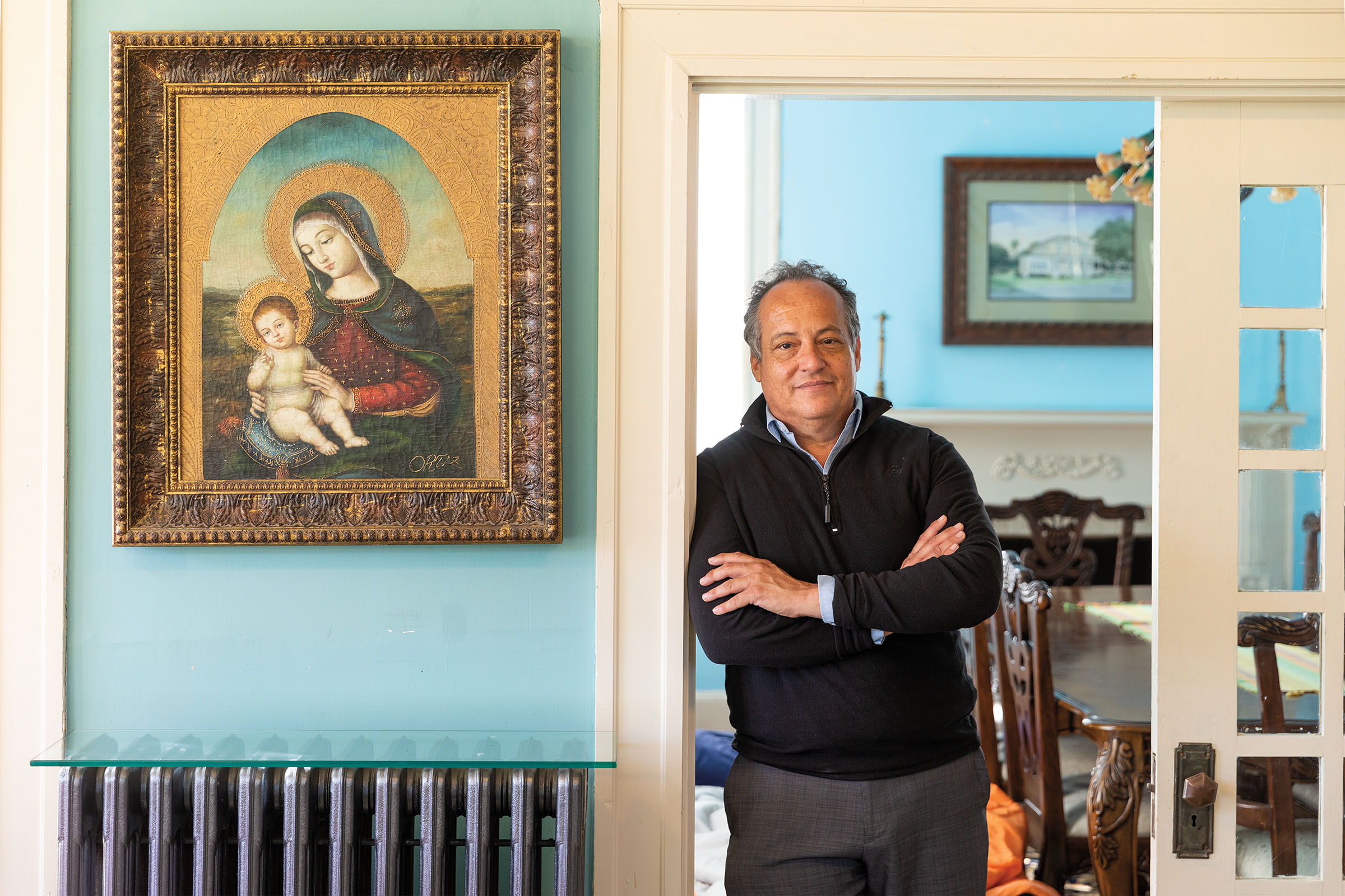 A man leans in the doorway inside of a historic home next to a religious painting on a turquoise wall