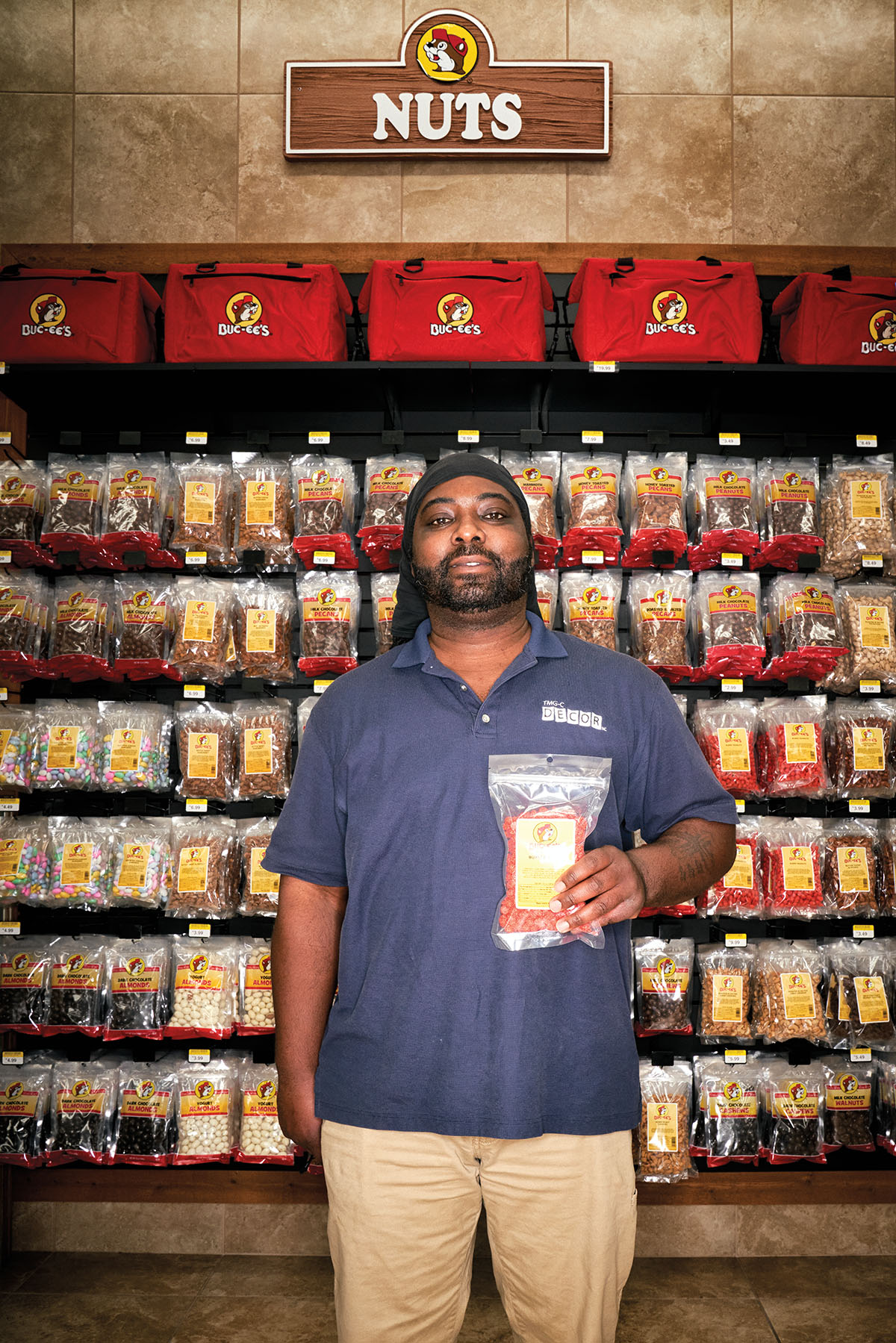 A man holds a bag of snacks in front of the large wall of hanging items at Buc-ees labeled "Nuts"