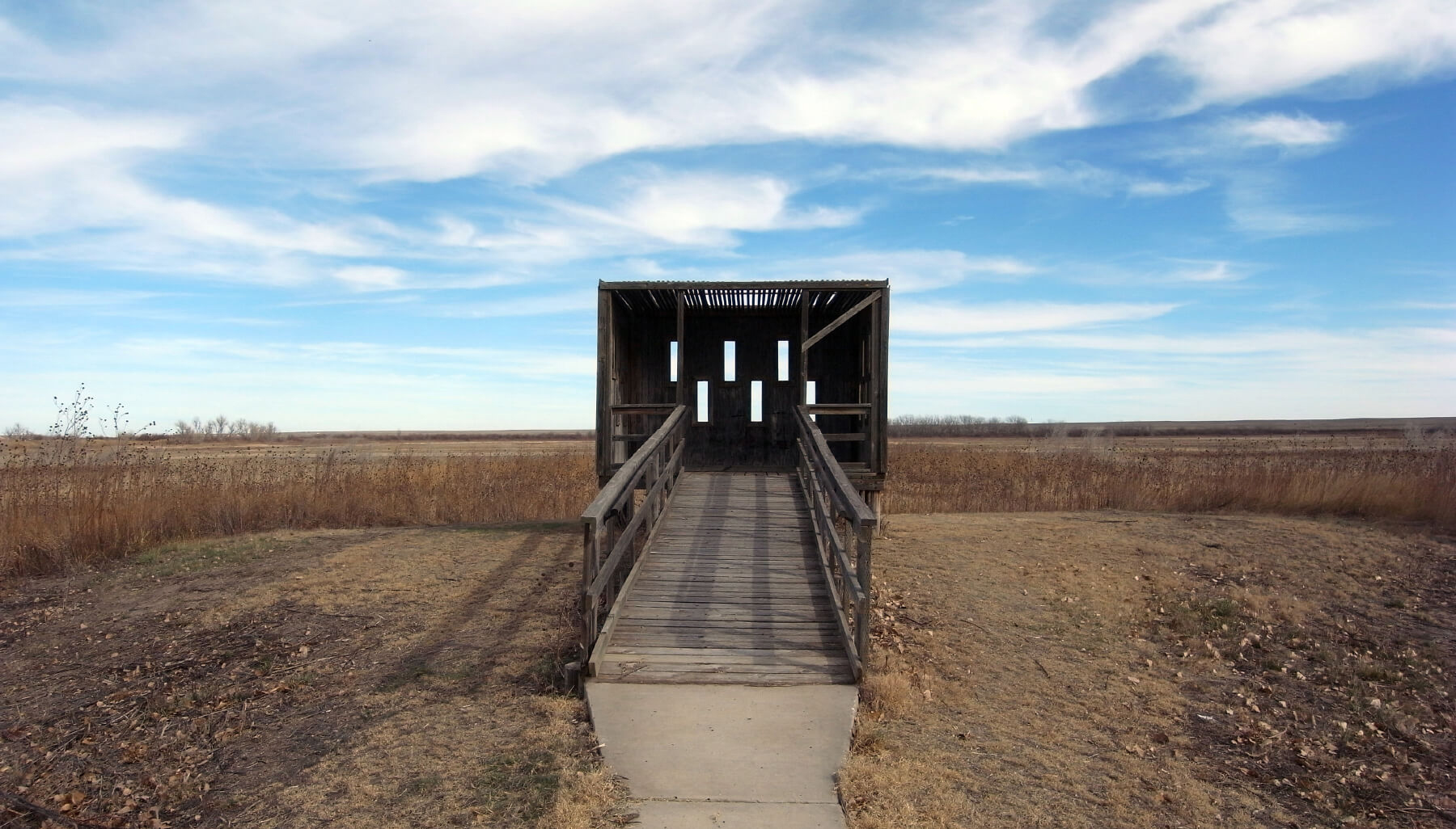 A wooden bird blind in a large brown field under a blue sky with clouds
