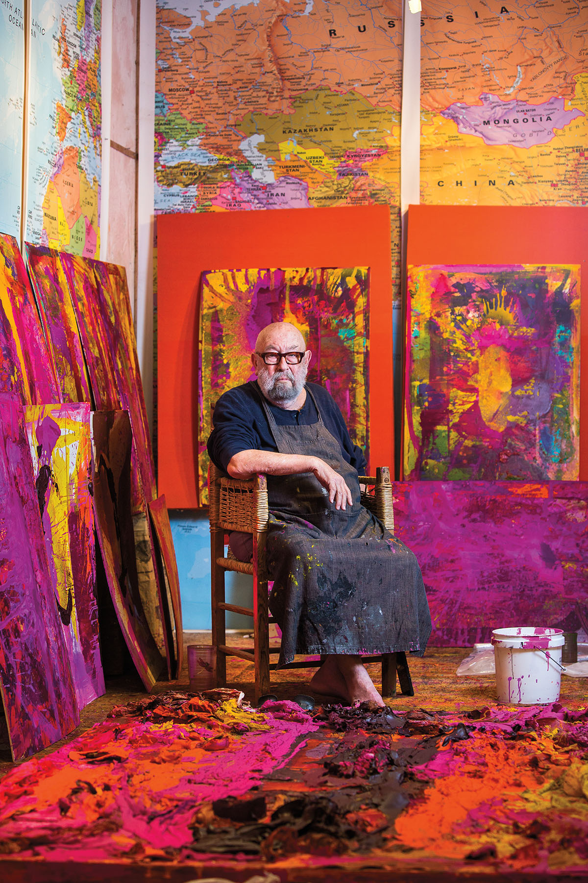 A man in a paint-spattered apron sits barefoot in a brightly-colored studio filled with pink, yellow and red paint