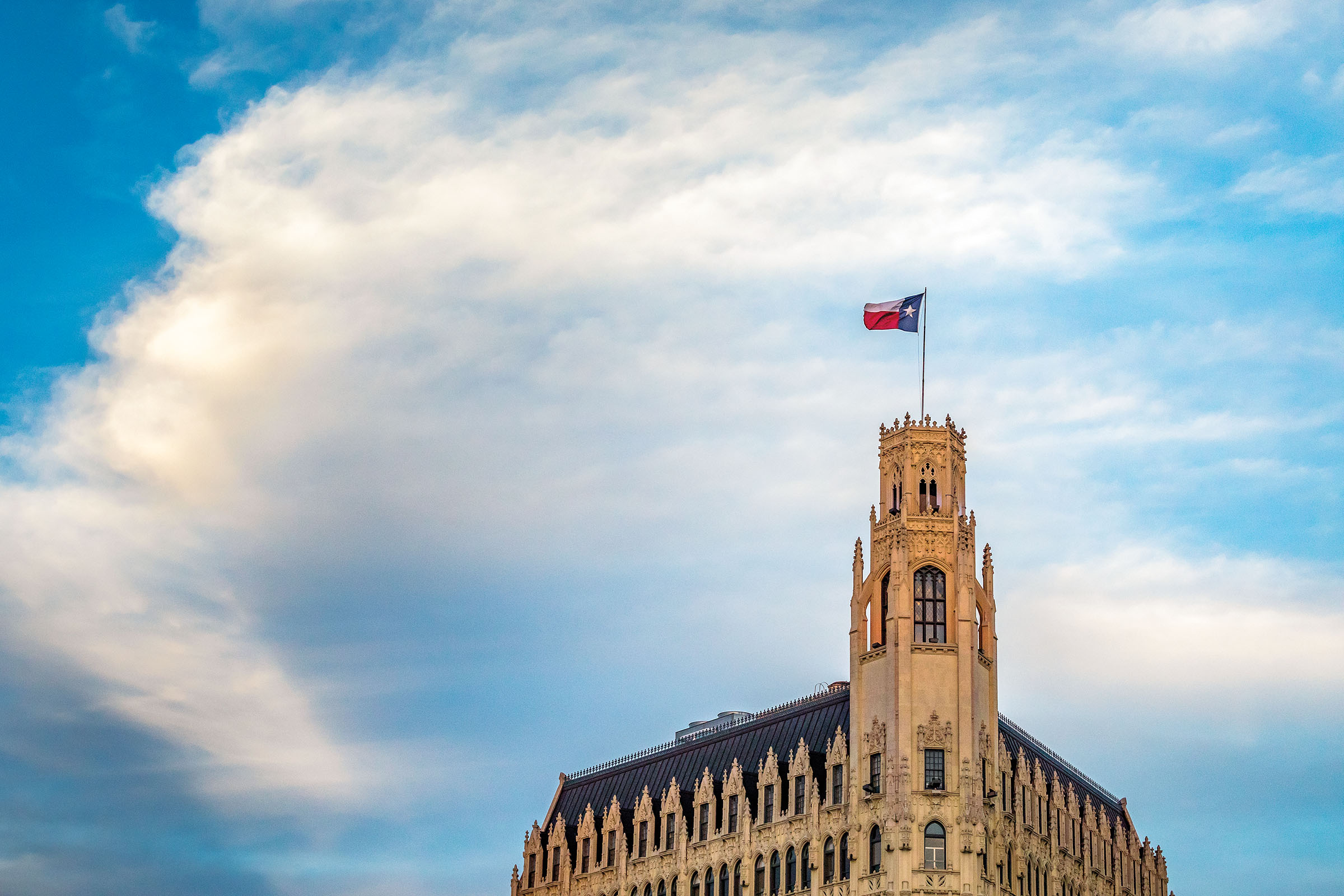 The top of an old building adorned with a Texas flag in front of a blue sky with clouds