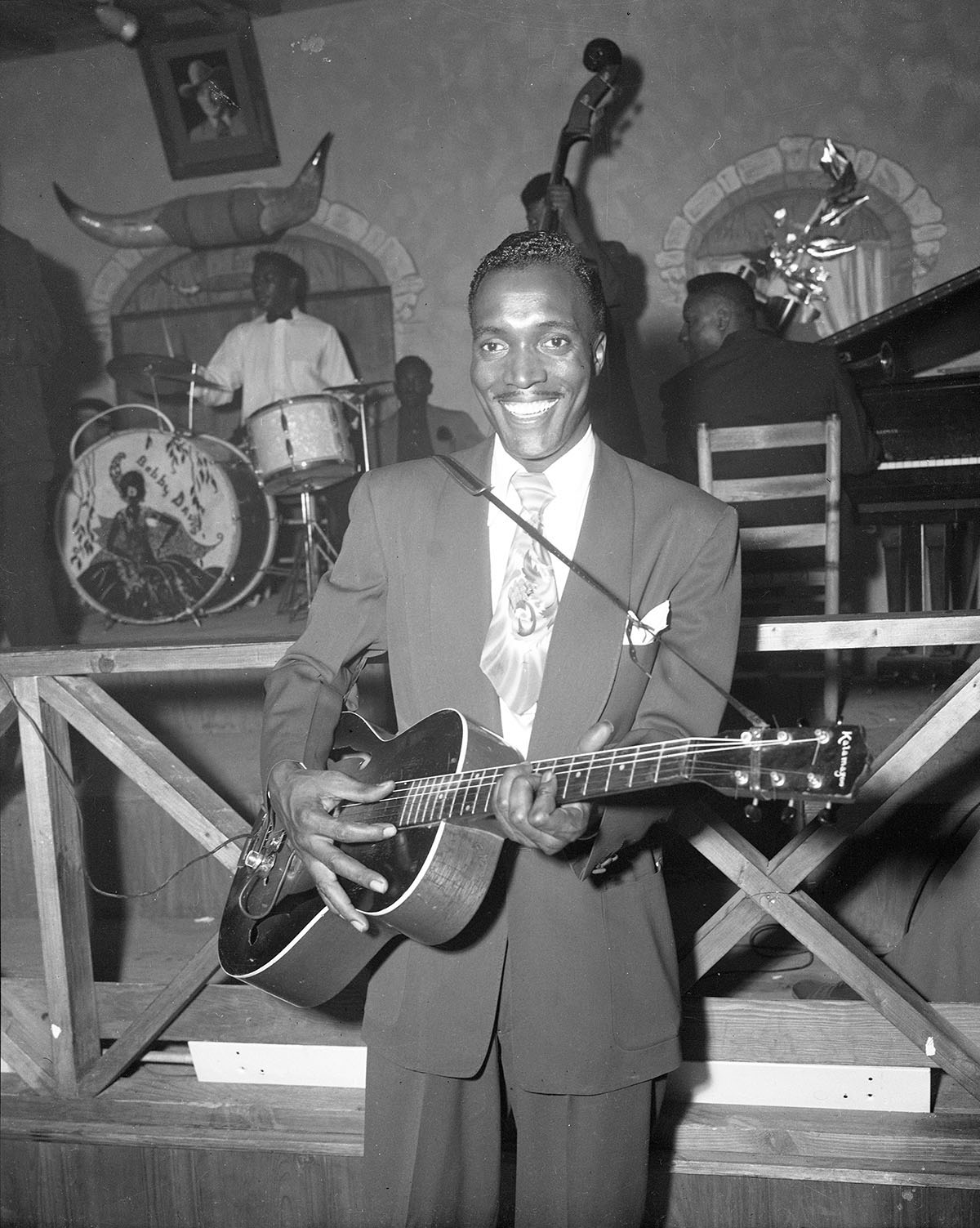 A man in a suit jacket holds an acoustic guitar