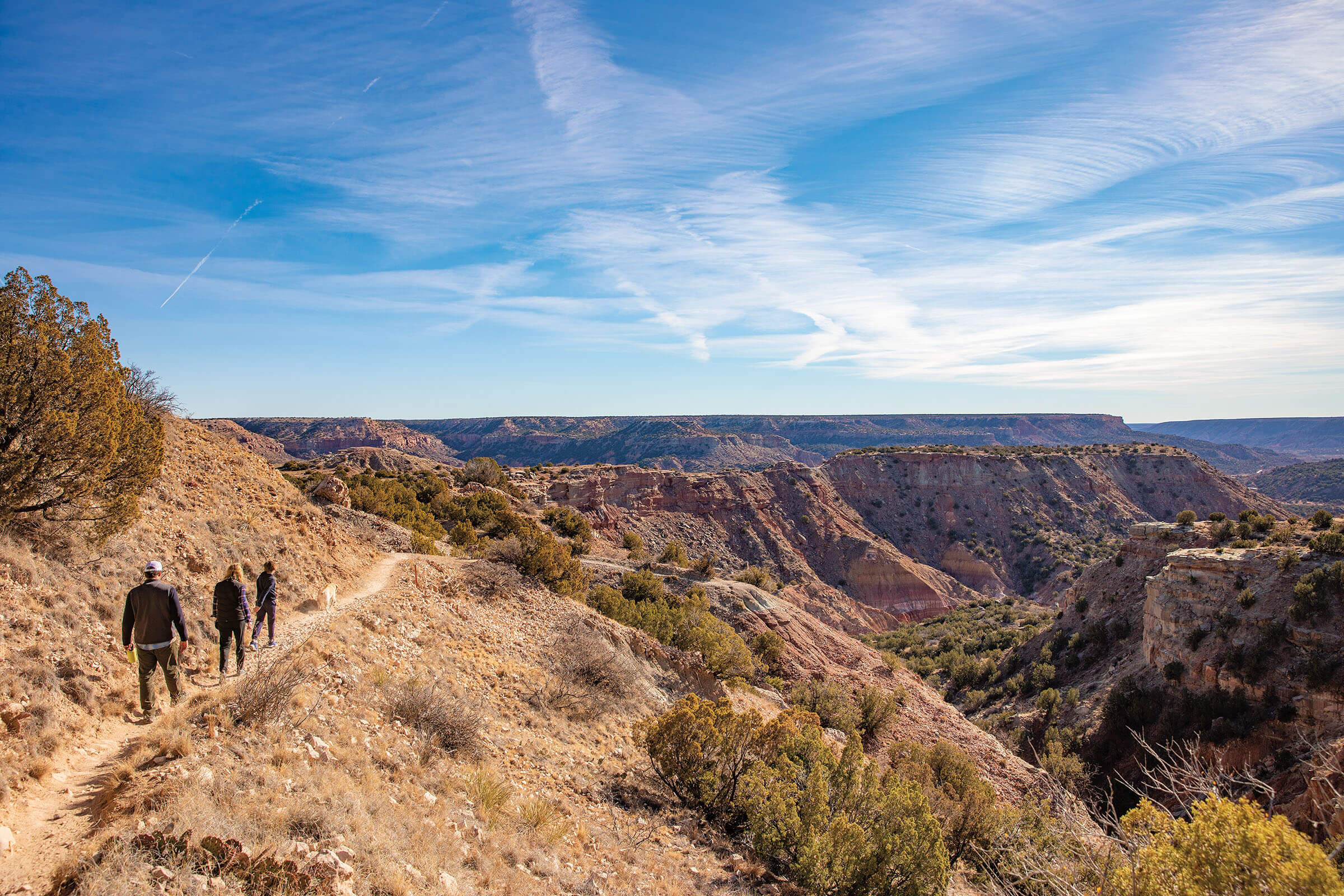 A wide view of a small group of hikers on a dirt trail among canyons under blue sky