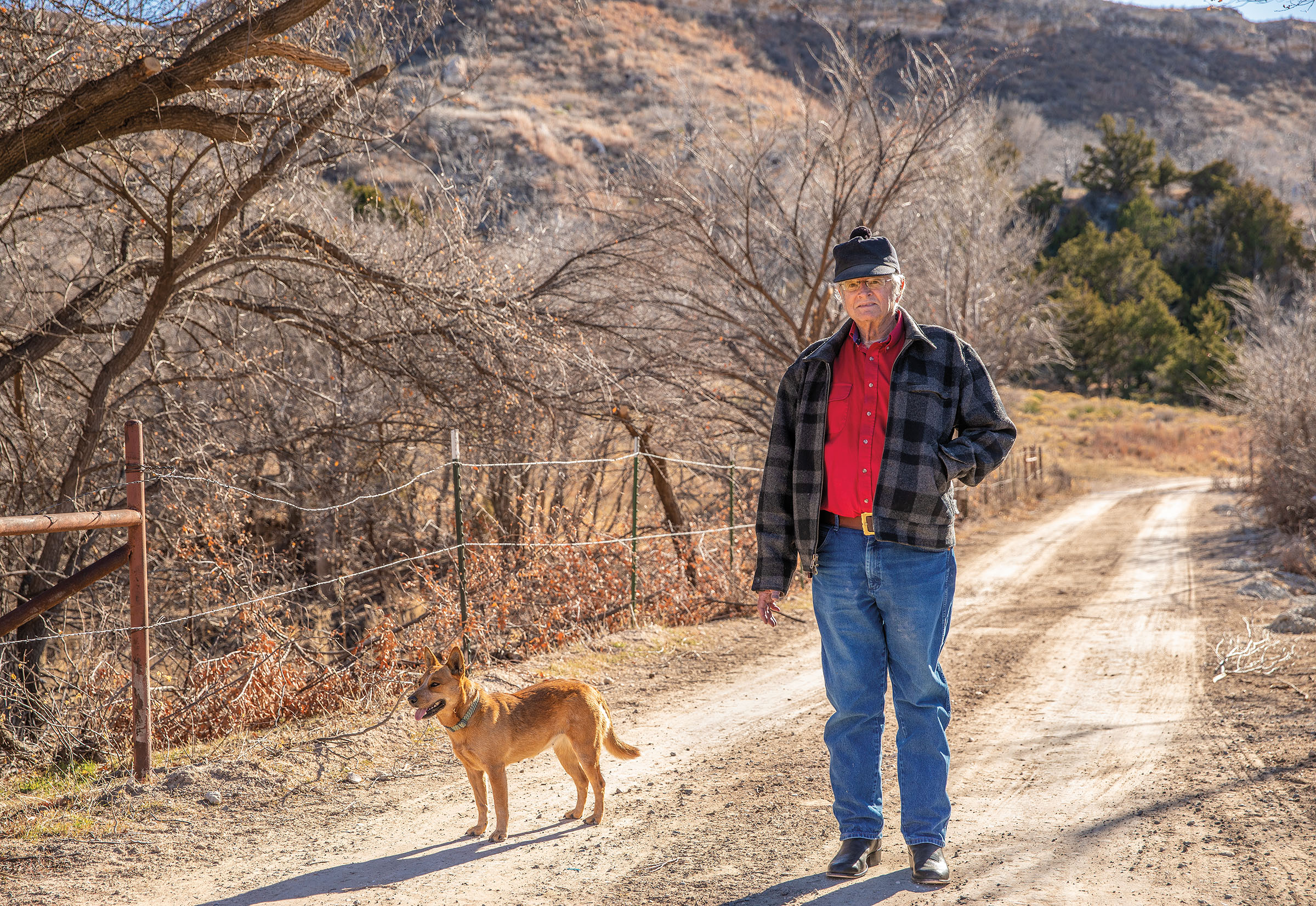 A man in blue jeans and a red shirt walks down a dirt path next to his tan dog