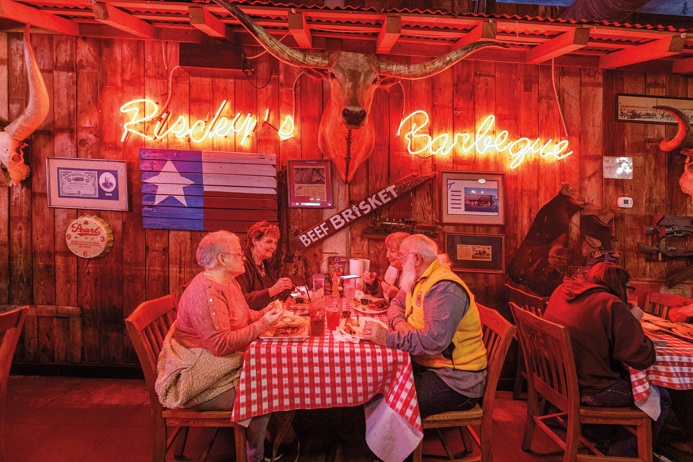 A group of diners eat at a red and white checkered picnic table under a neon sign reading "Riscky's Barbecue" next to a Texas flag