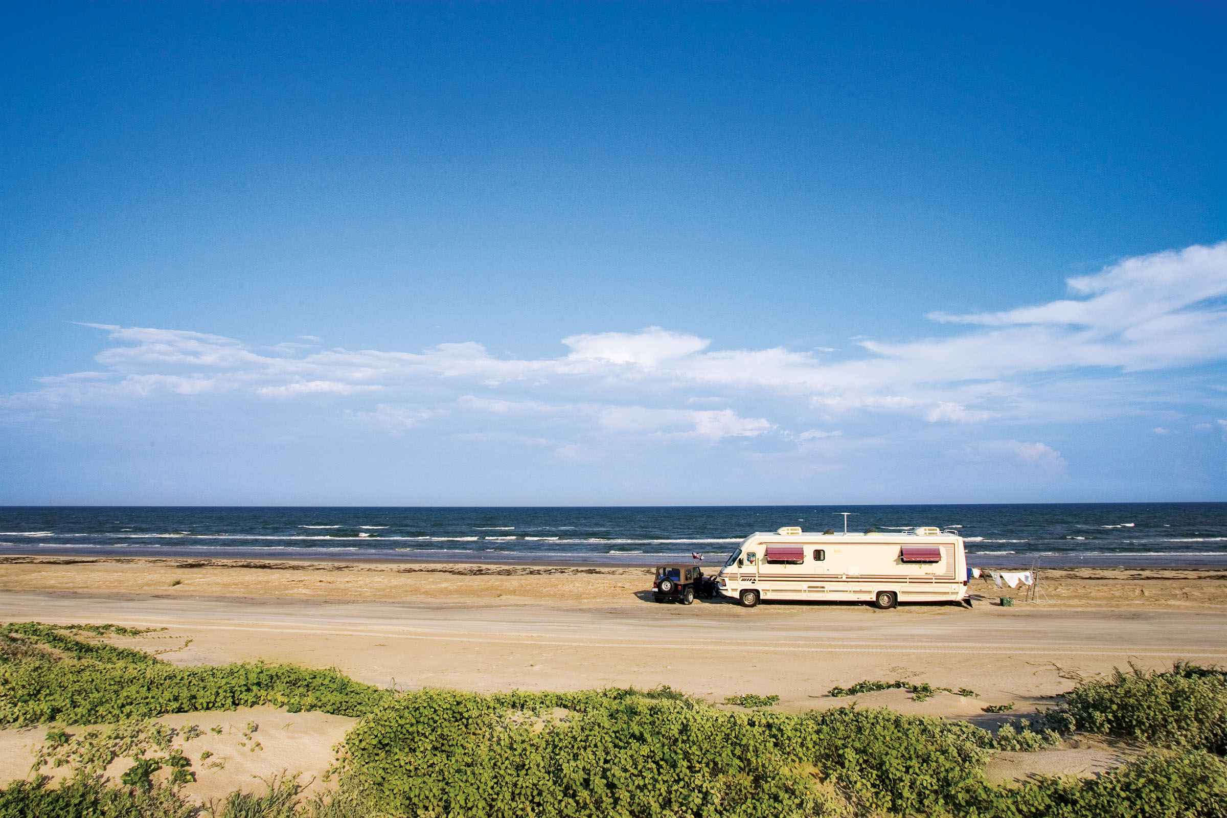 An RV is pulled behind a truck along a sandy beach