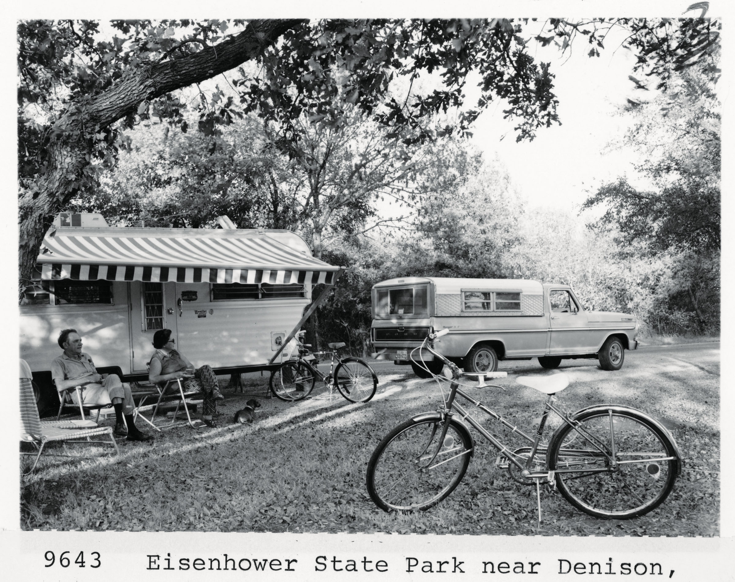 A historic photo of a camper parked at a ampsite with two people lounging in front