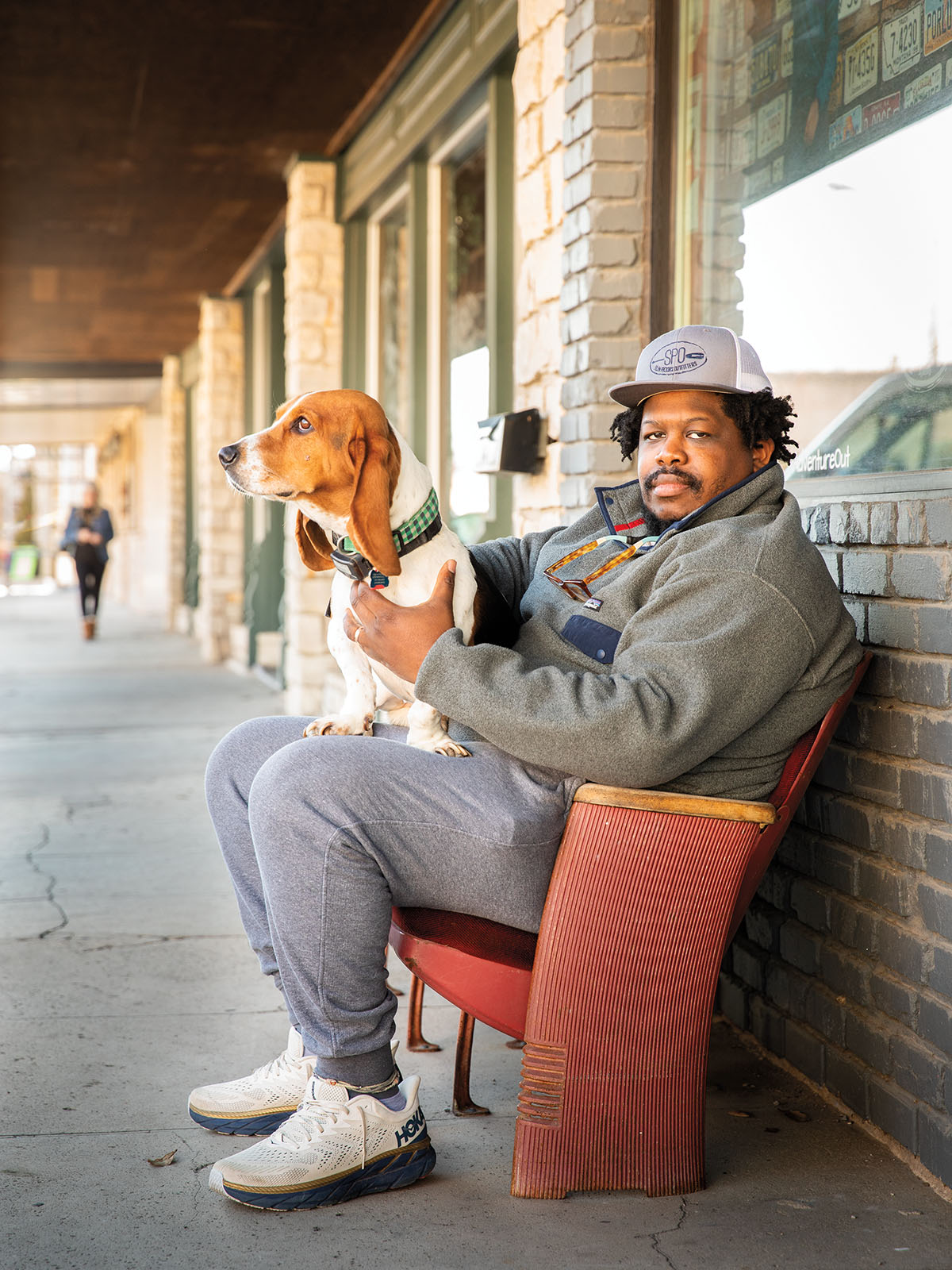 A man in a fleece jacket and gray pants sits in a chair with a dog in his lap