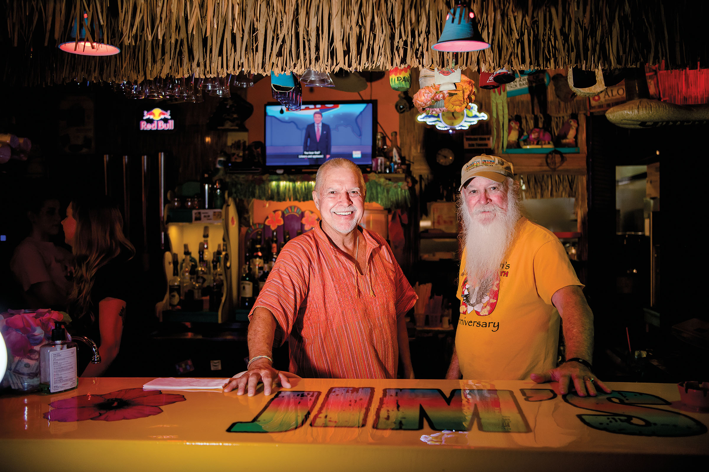 Two men smile behind a warmy-lit bar with a yellow bartop reading "Jim's"