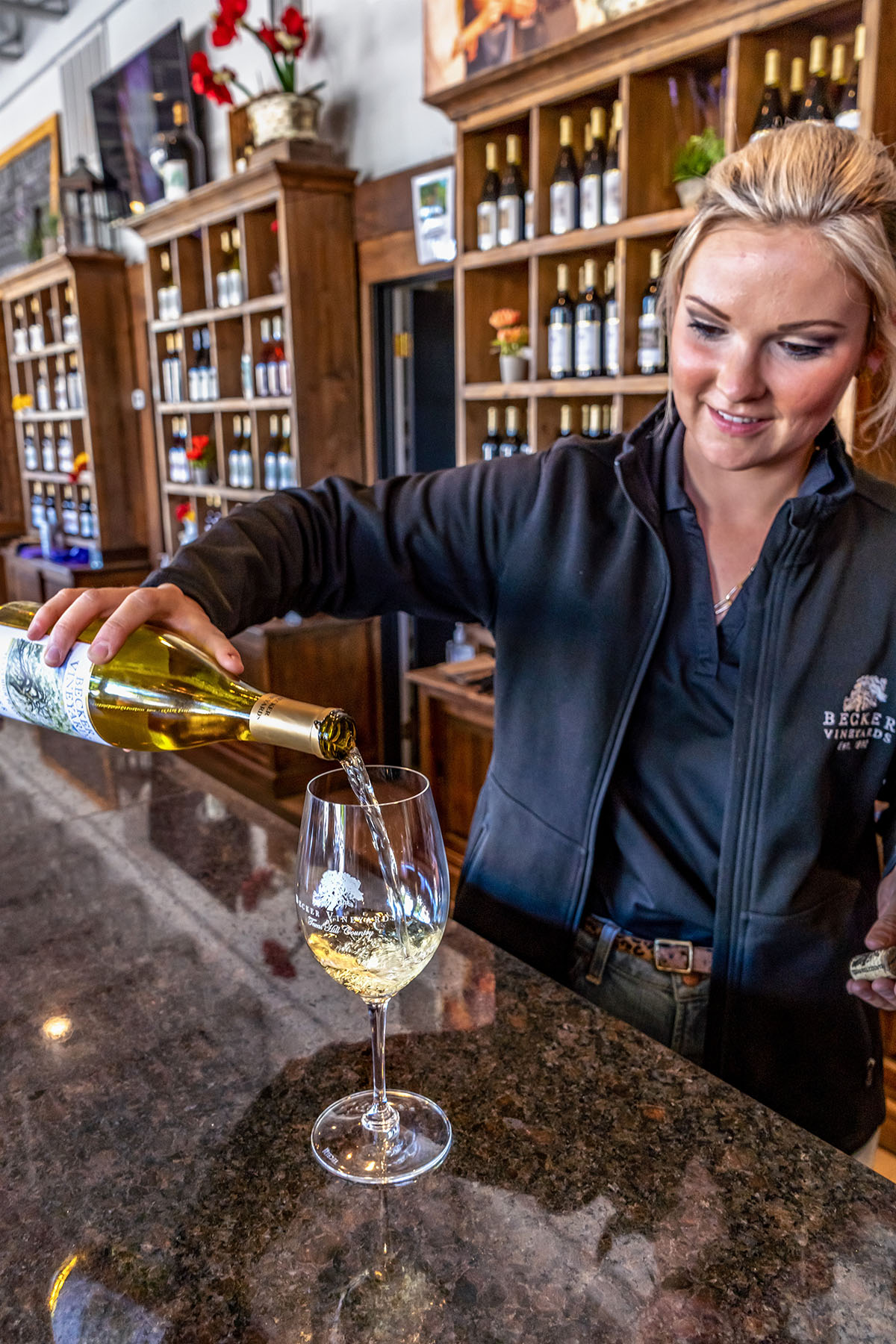 A woman in a black shirt pours white wine into a glass reading "Becker Vineyards"