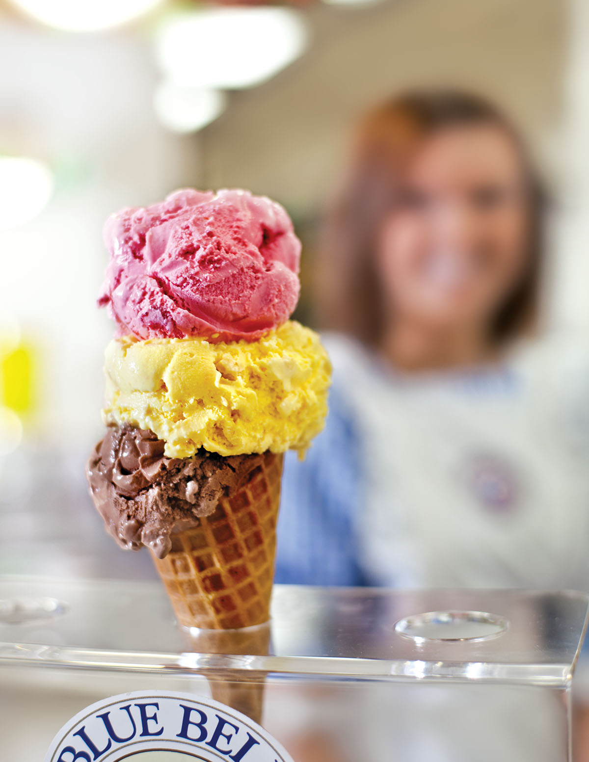 A pink, yellow and brown scoop of ice cream perched in a waffle cone with a woman smiling in the background