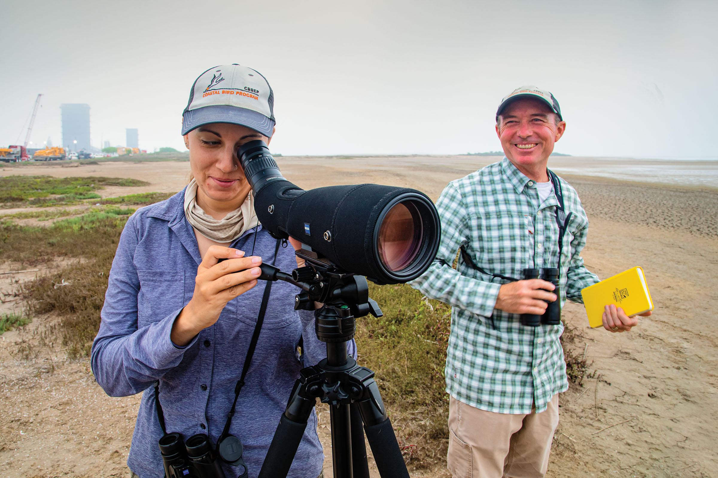 Two people stand with a telescope, binoculars and a small book on the shoreline