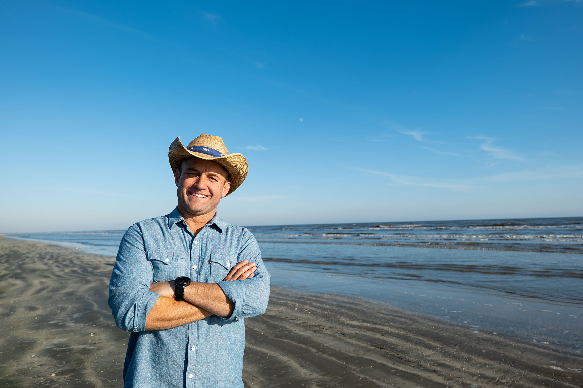 A man in a cowboy hat stands with his arms crossed by the sea