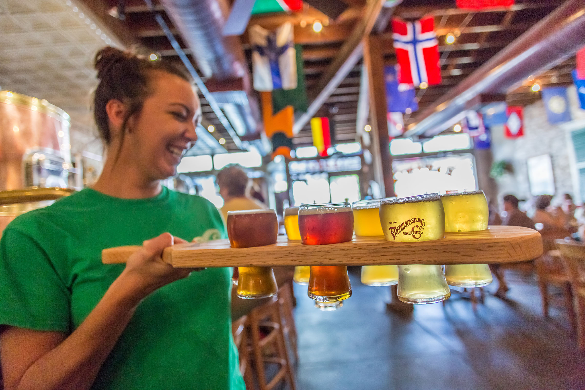 A woman in a green shirt holds a wooden tray full of beers