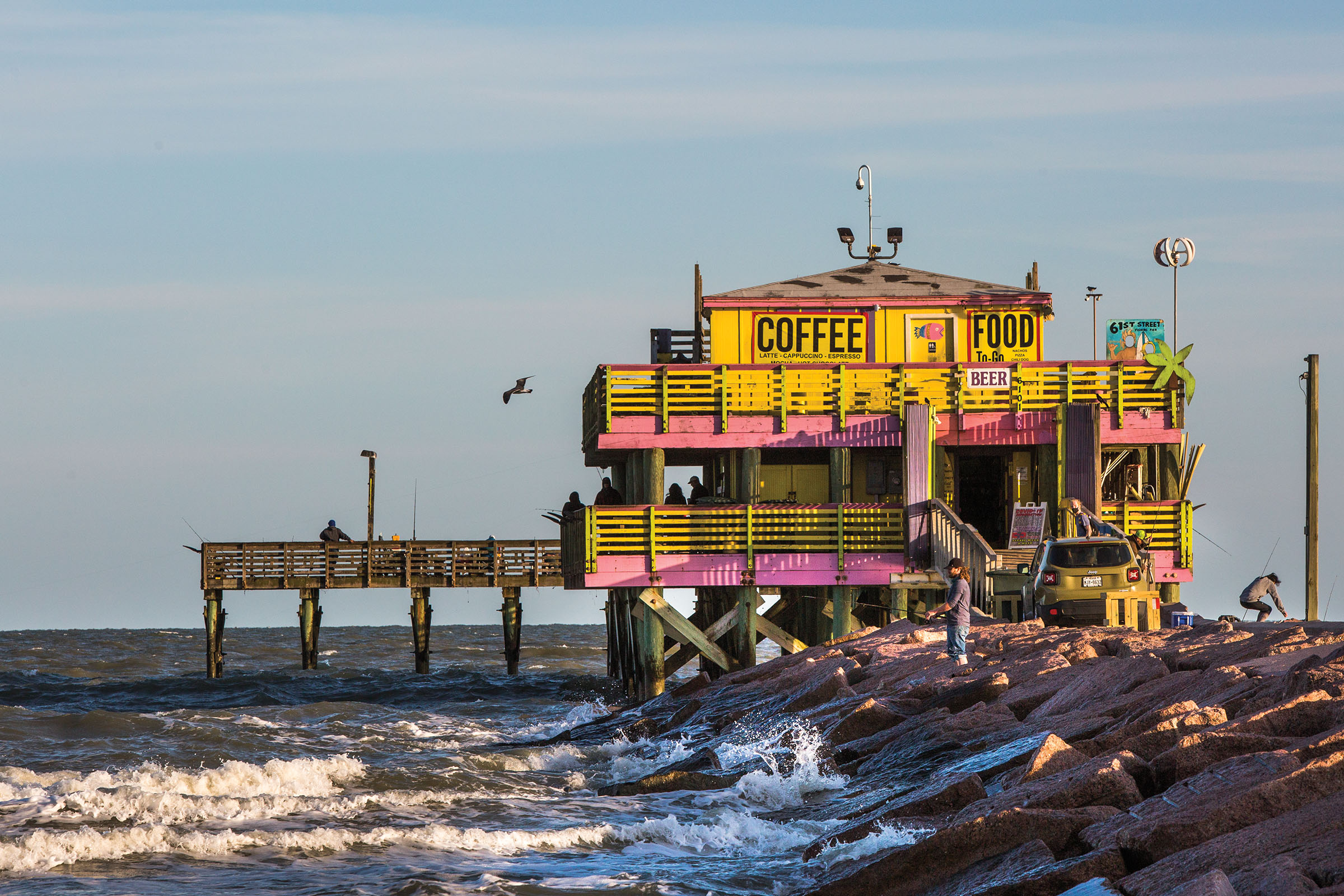 A brightly colored, pink and yellow building with a pier sits atop crashing blue waves