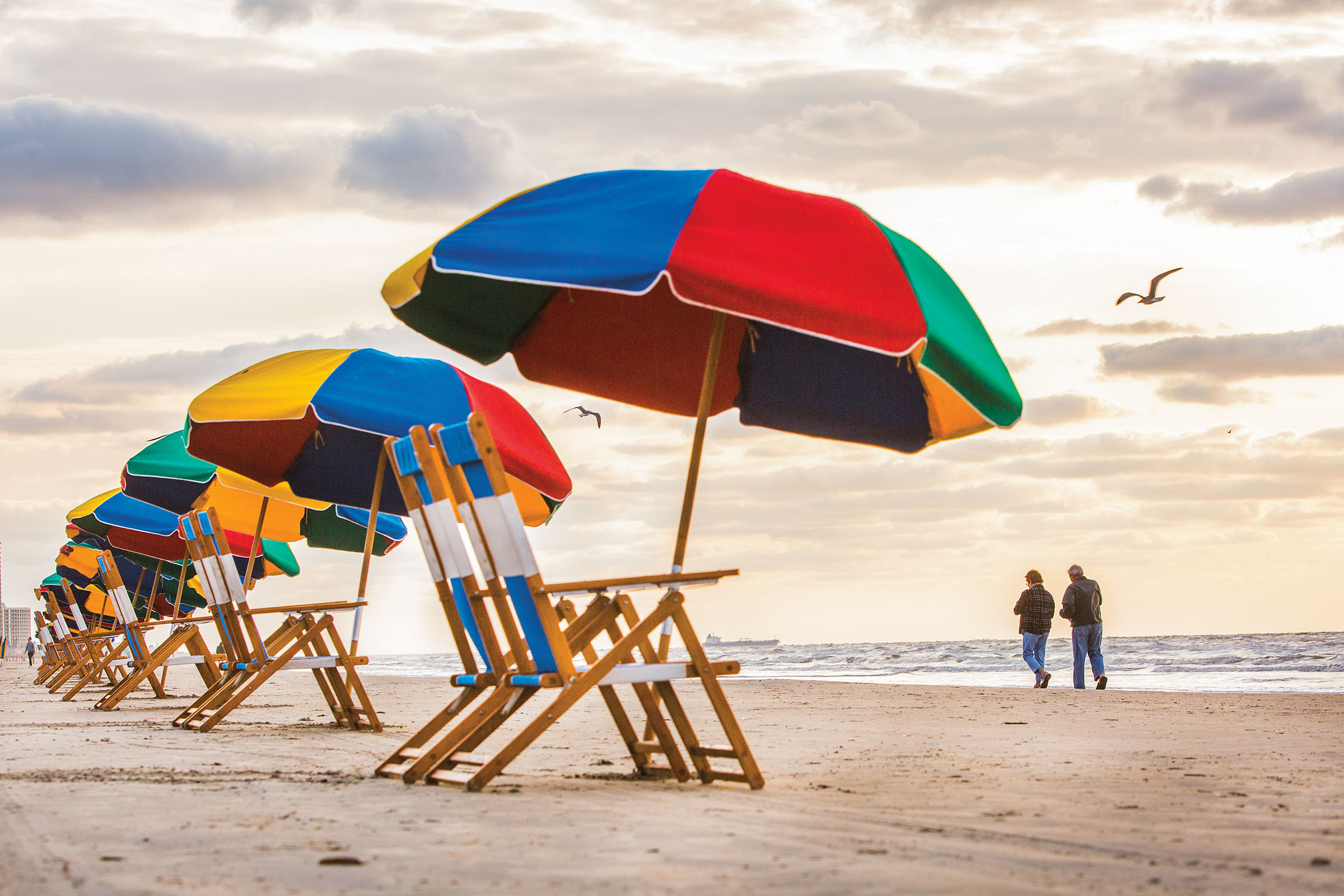 Brightly colored beach umbrellas stand in the sand above beach chairs while a couple walks in the sand